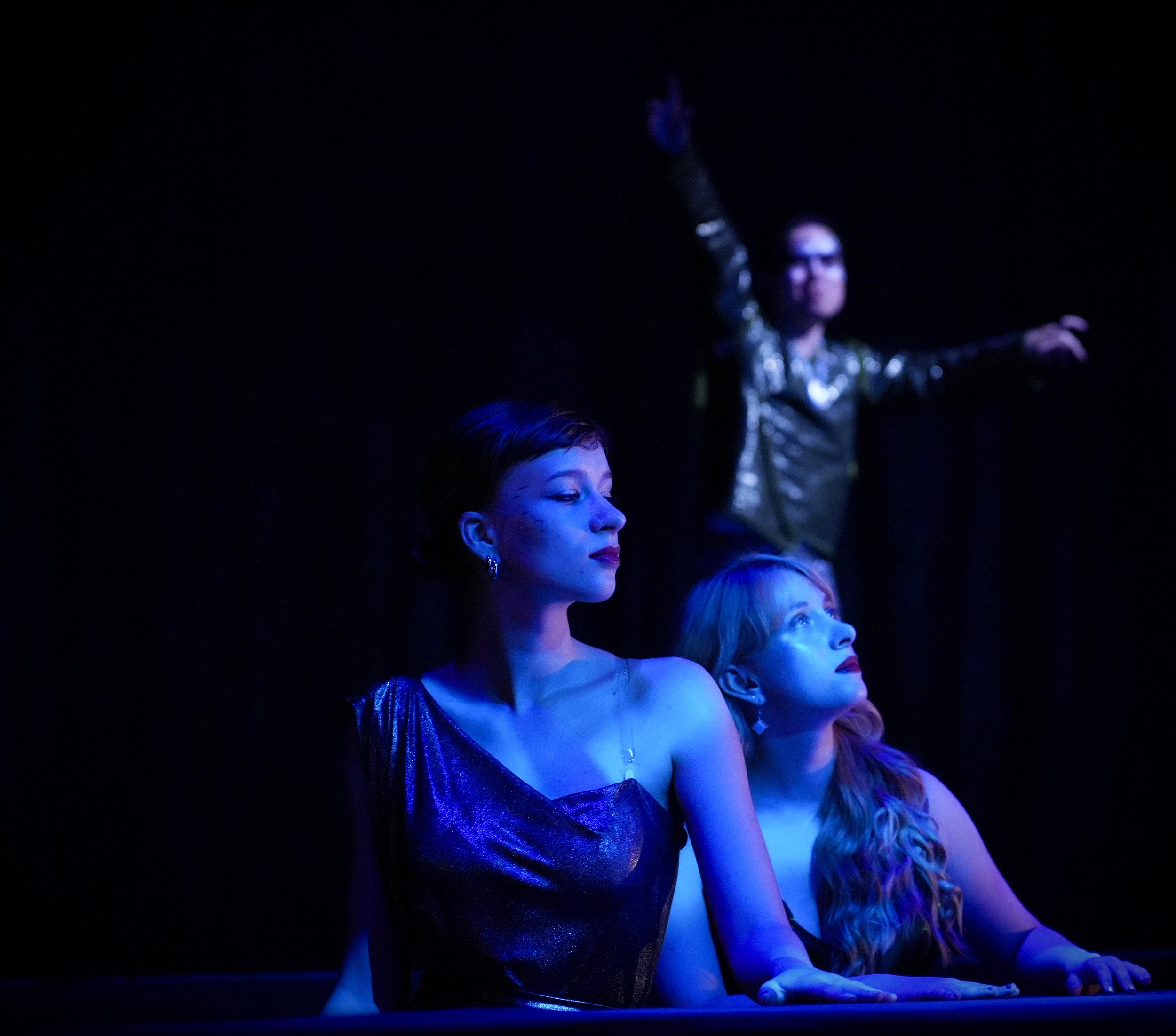 Three women with blue lighting, two are seated and looking to the left, one standing in the background with arms raised.