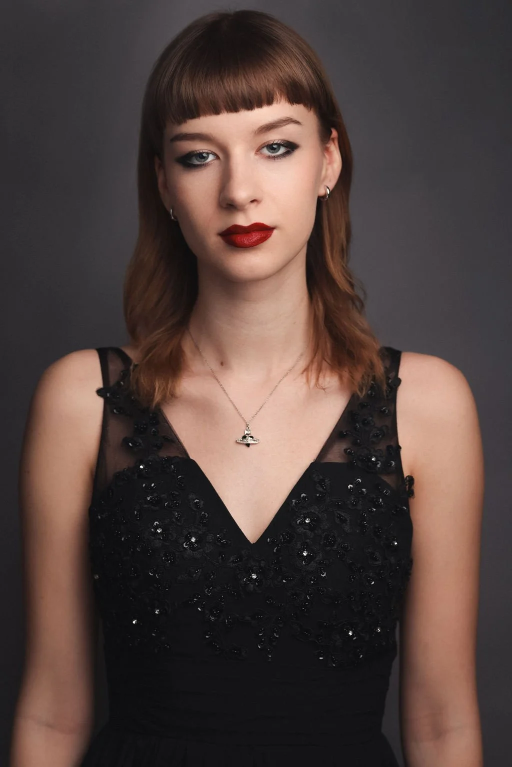 A woman with brown hair and blunt bangs, wearing a black sleeveless dress with beaded embellishments and a silver necklace featuring a UFO pendant, standing against a gray background.