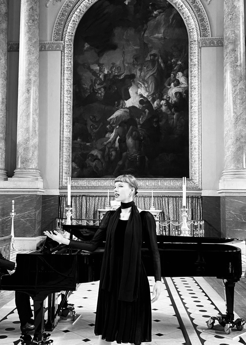A woman singing at a grand piano in a historic concert hall with ornate decorations, large painting, and candles.