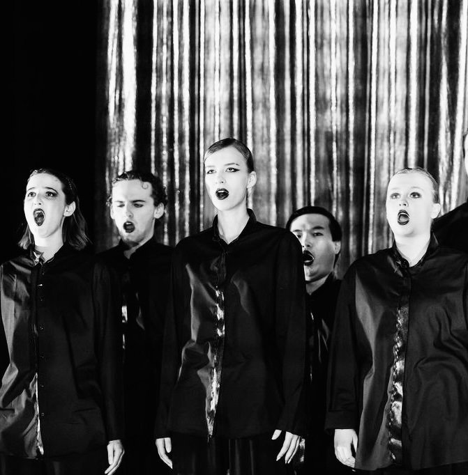 Black and white photo of five female singers standing in front of a curtain, singing with open mouths.