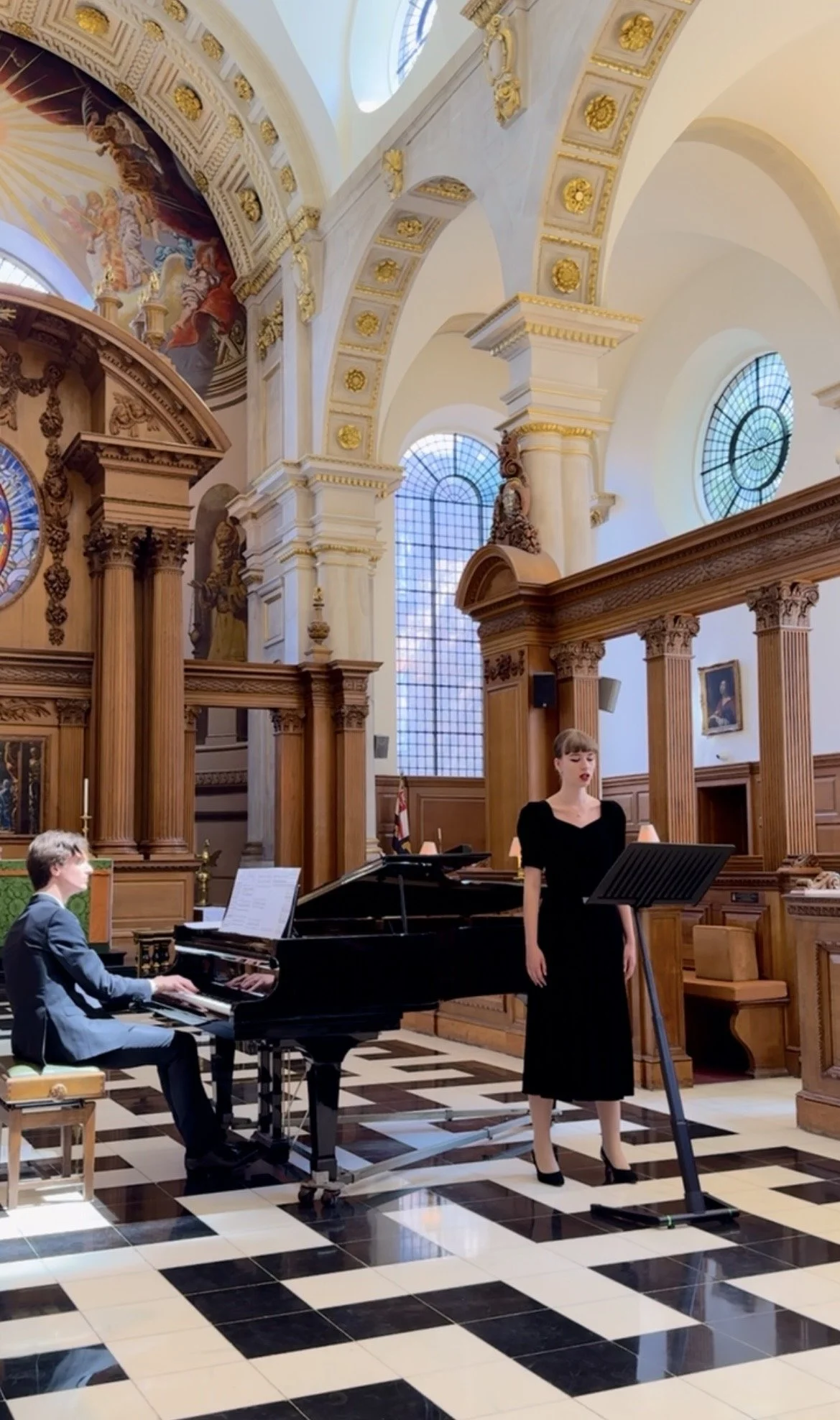 A piano and a singer performing inside a grand, ornately decorated church with gold accents, large stained glass windows, and a checkered black and white floor.