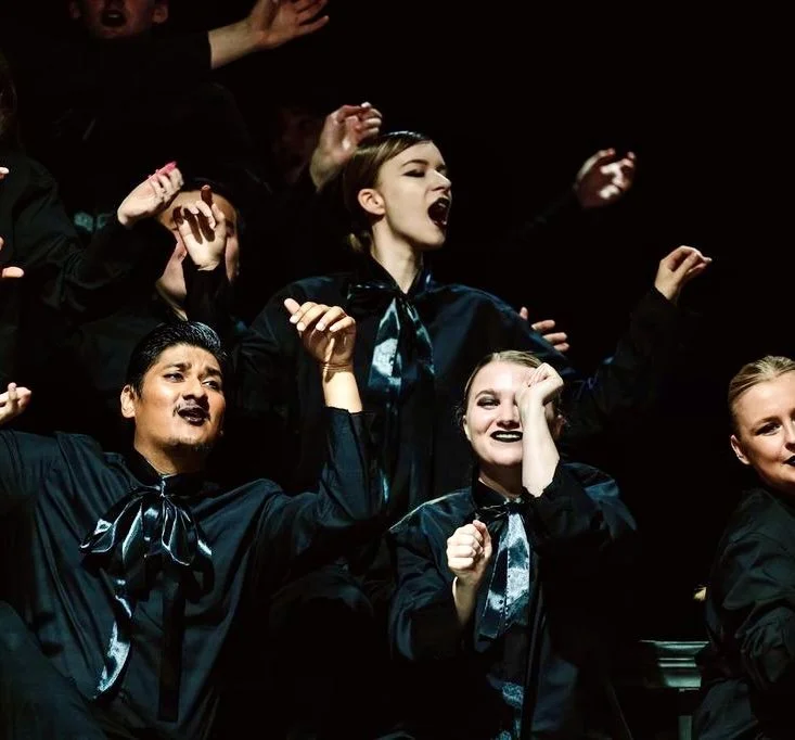 Group of young performers singing passionately, dressed in black costumes with shiny black scarves and makeup, stage lighting highlighting their faces.