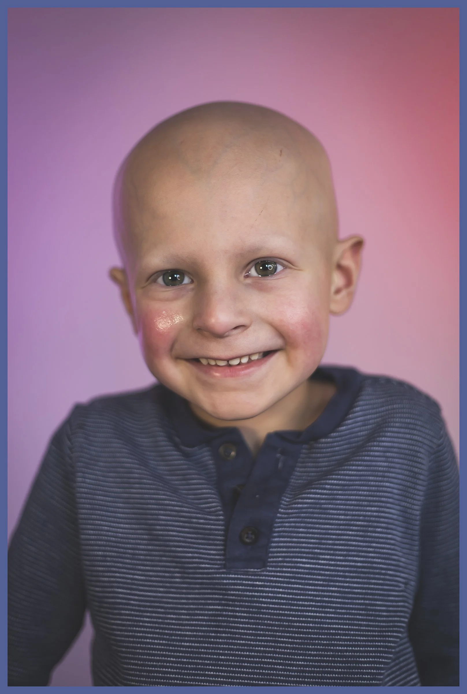 Close-up portrait of a smiling young boy with a shaved head, wearing a dark blue striped shirt, against a pink and purple gradient background.