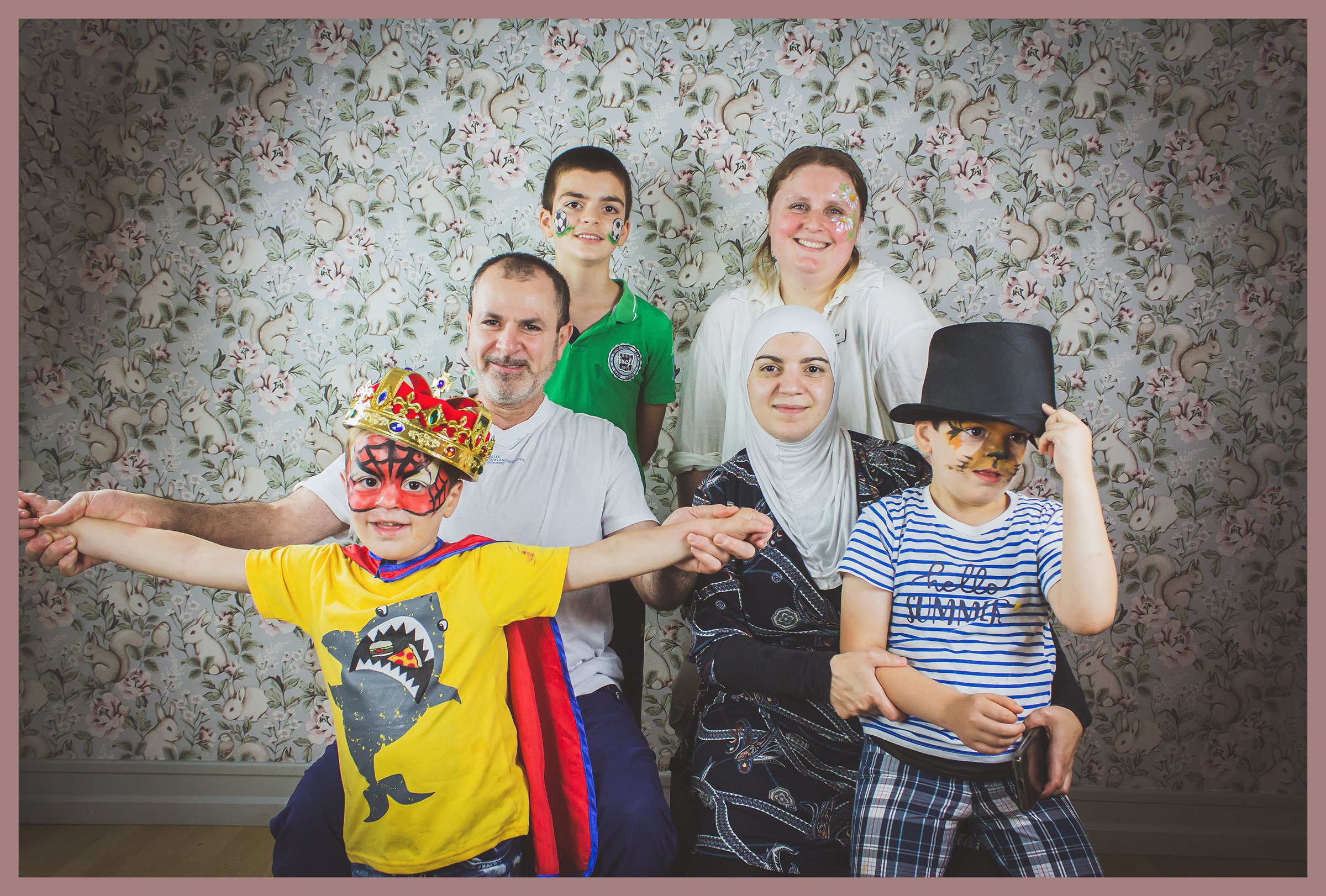 A group of six people, including children and adults, dressed in costumes or casual clothes, posing in front of a floral wallpaper background. Children are wearing face paint, and one is dressed as a tiger with a crown, while another has a striped sh