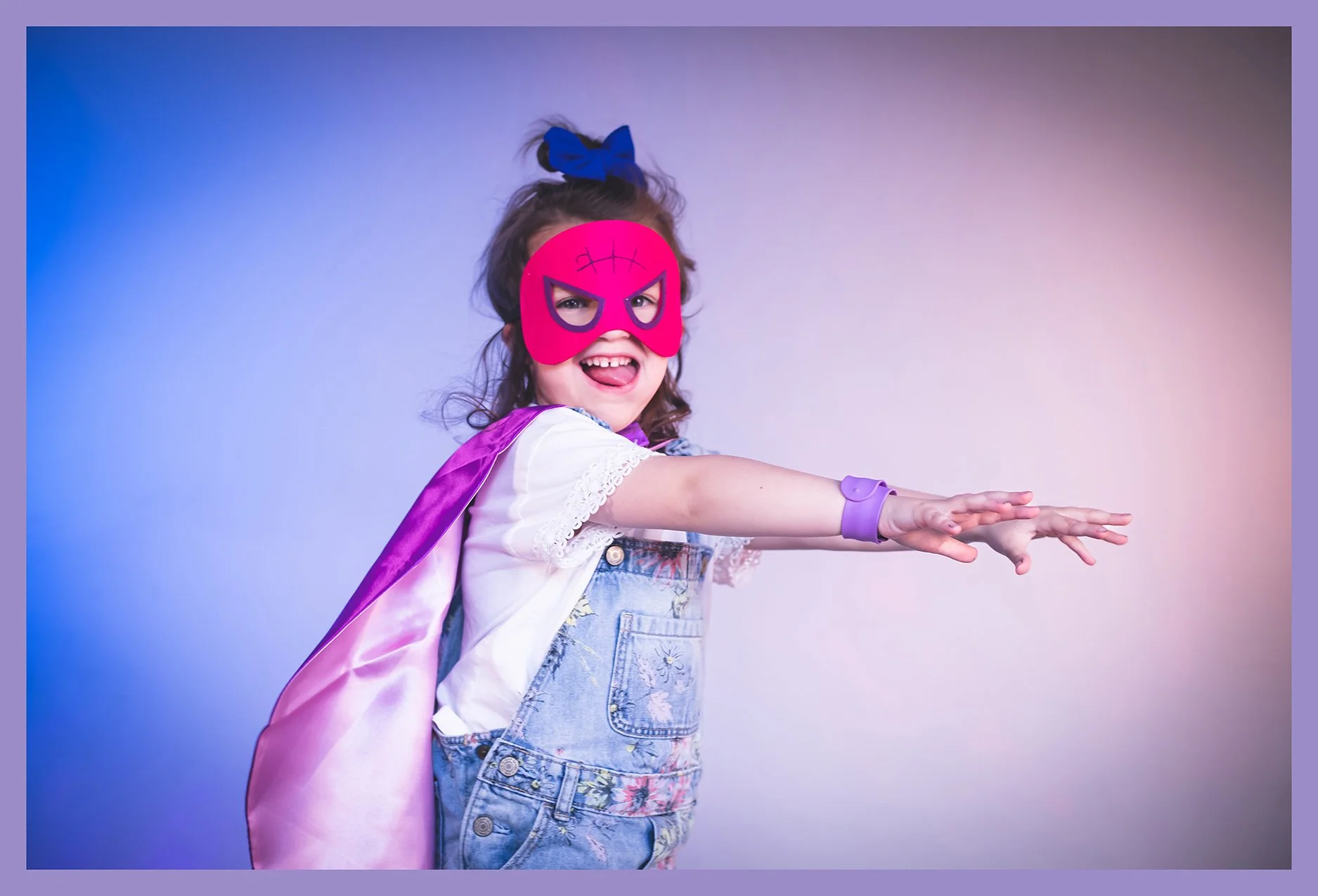 Young girl dressed as a superhero, wearing a pink mask, purple cape, denim overalls, and a white shirt, with her arms extended forward as if flying or reaching out, smiling with tongue out, and posing against a gradient background.
