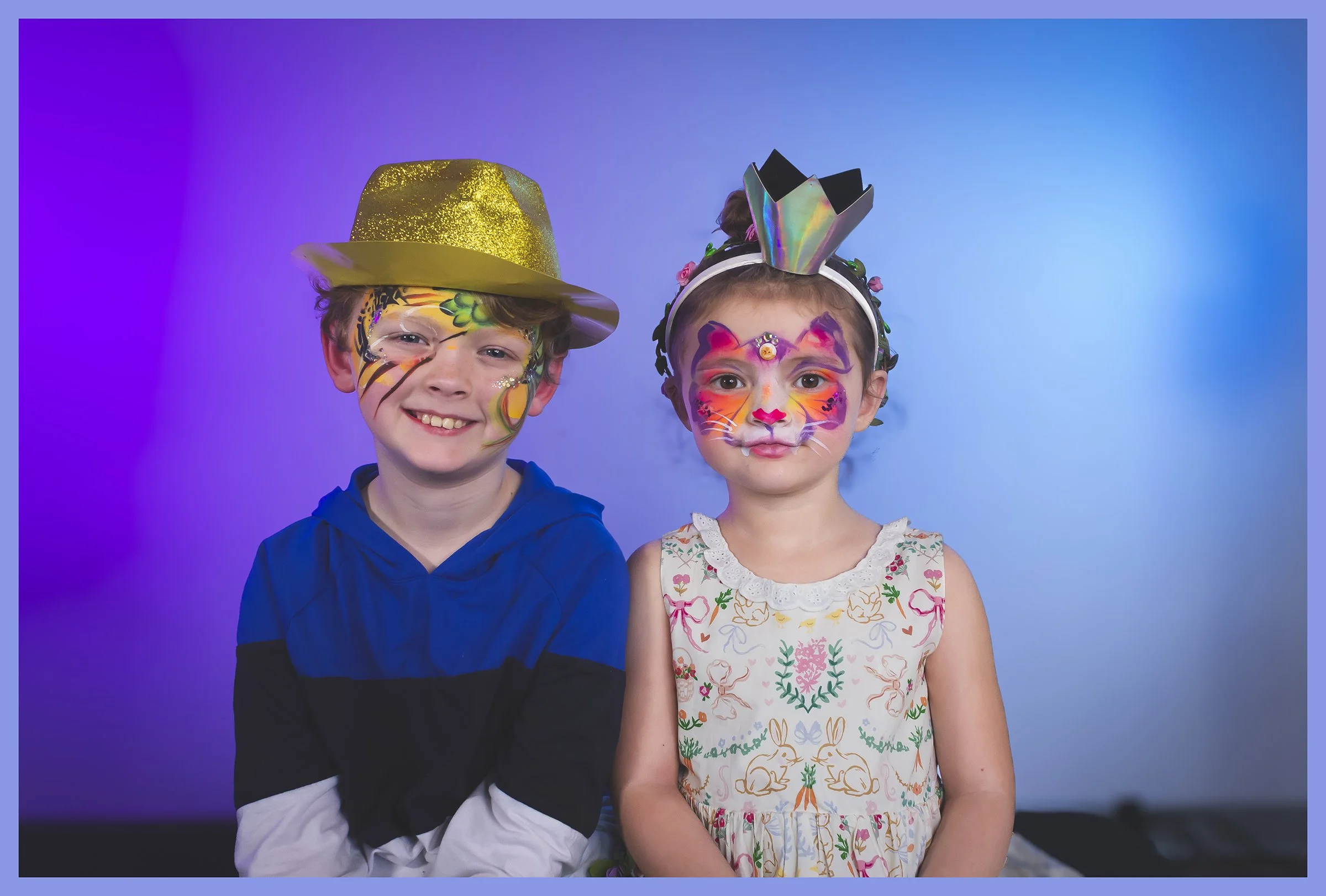 Two children with colorful face paint and costumes posing against a blue and purple gradient background.
