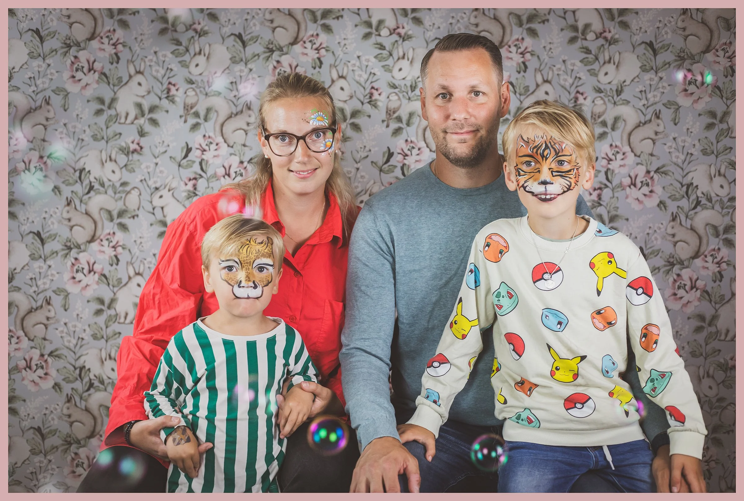 A family of four posing in front of a floral cartoon rabbit wallpaper. The mother and one child have tiger face paint, and the other child has a lion face paint. The boy is wearing a Pikachu sweatshirt, and the girl is wearing a Pokémon-themed shirt.
