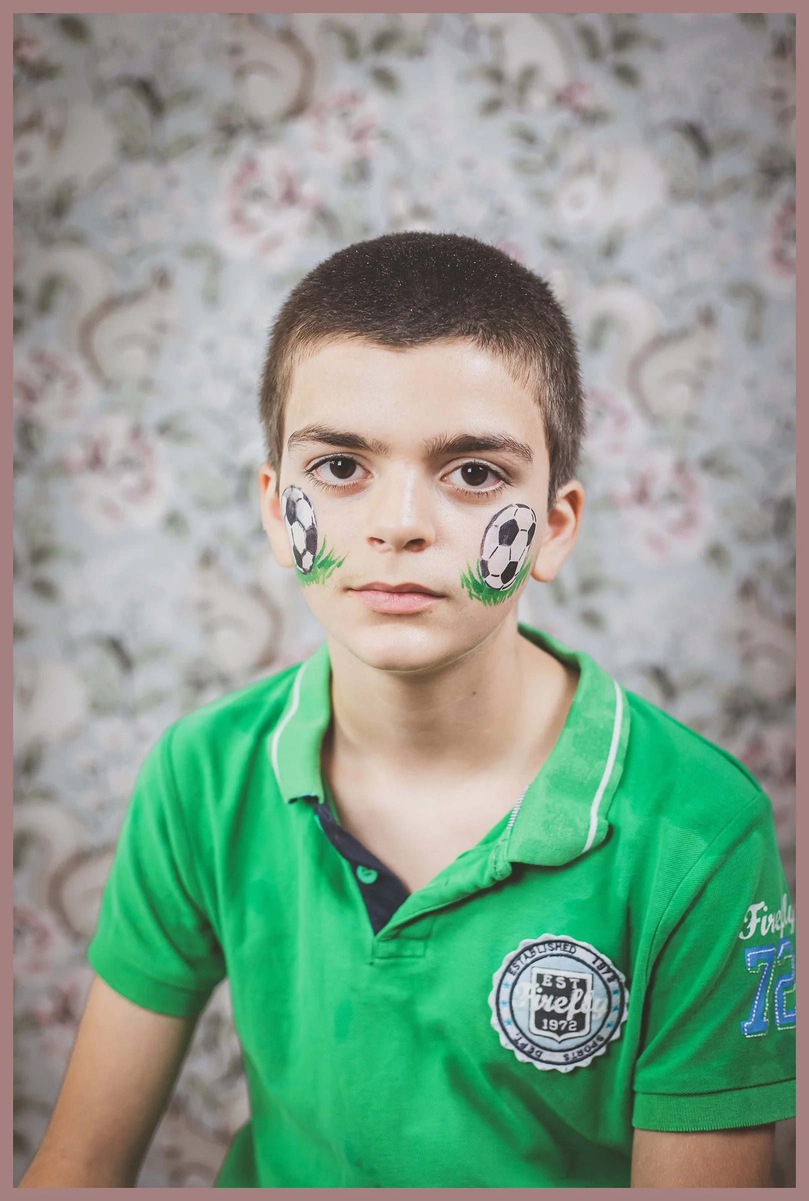 Portrait of a young boy with face paint of soccer balls and grass on his cheeks, wearing a green polo shirt, with a floral background.
