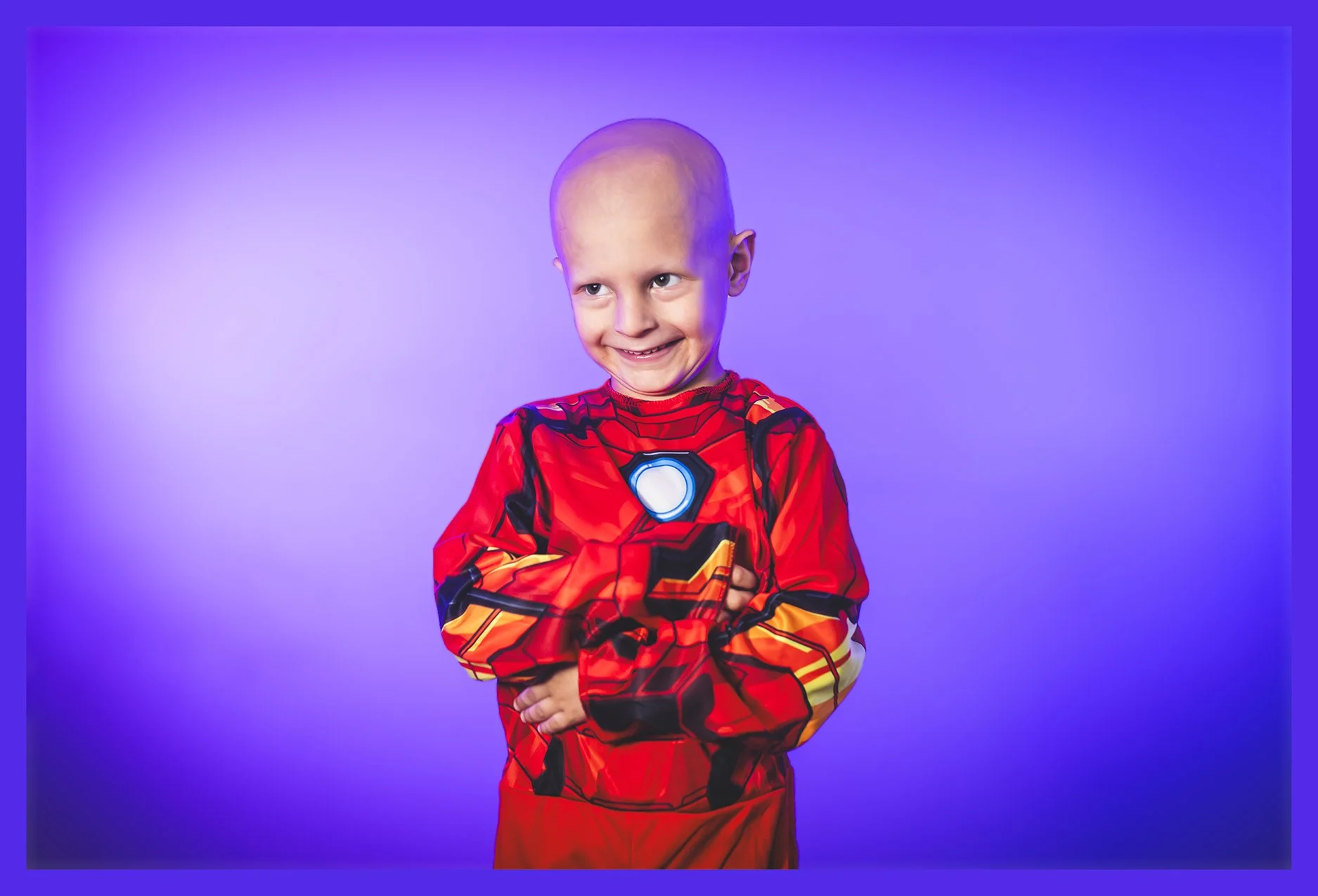 A young boy with a bald head wearing a red Iron Man costume, smiling mischievously, standing against a purple background.