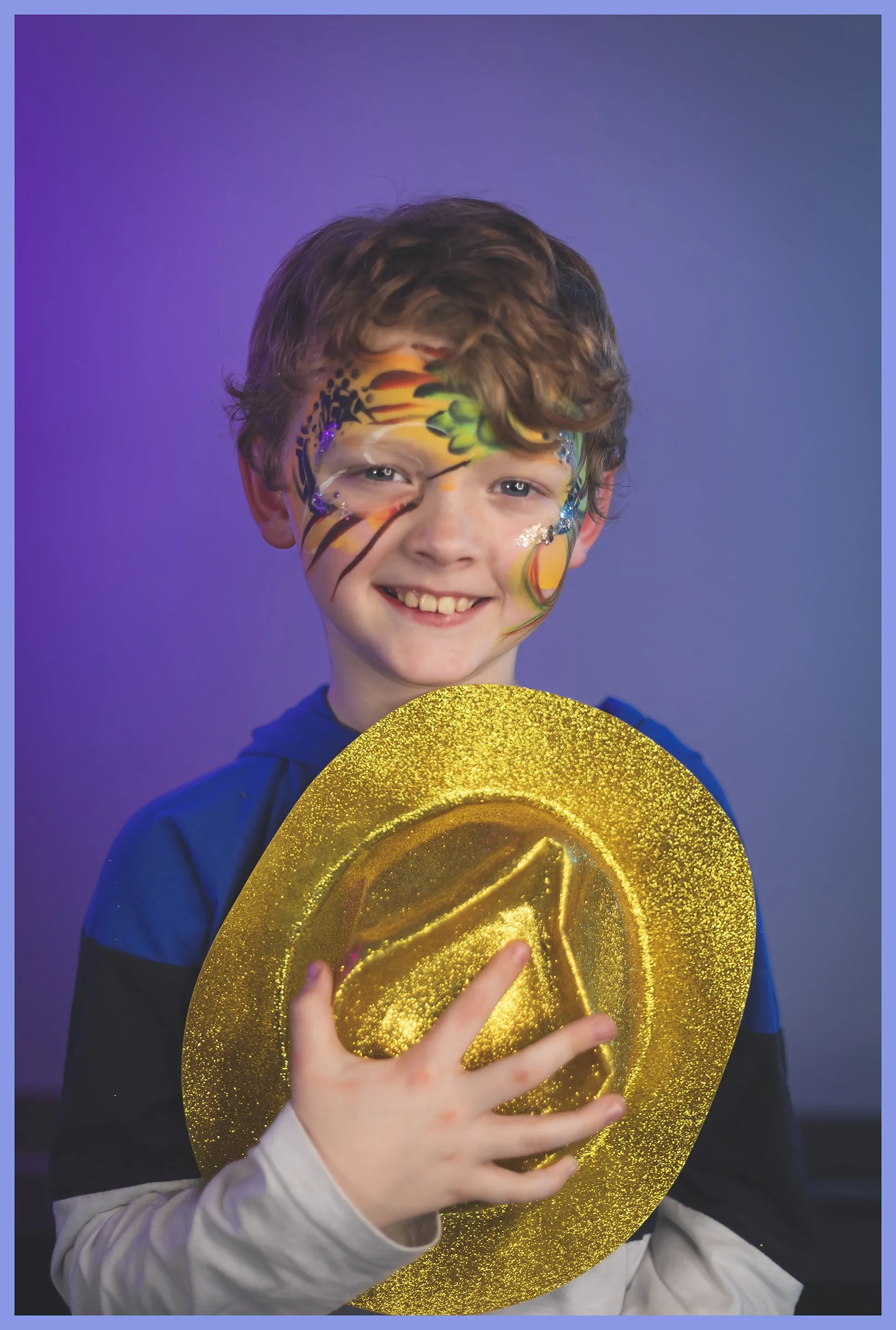 A smiling young boy with face paint resembling a butterfly, holding a shiny gold hat, against a purple background.