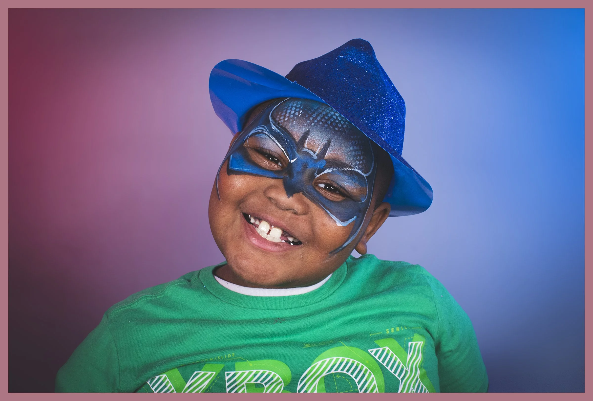 A young boy smiling with a missing tooth, wearing a green shirt and a blue wizard hat, with face paint resembling a superhero mask against a gradient pink and blue background.