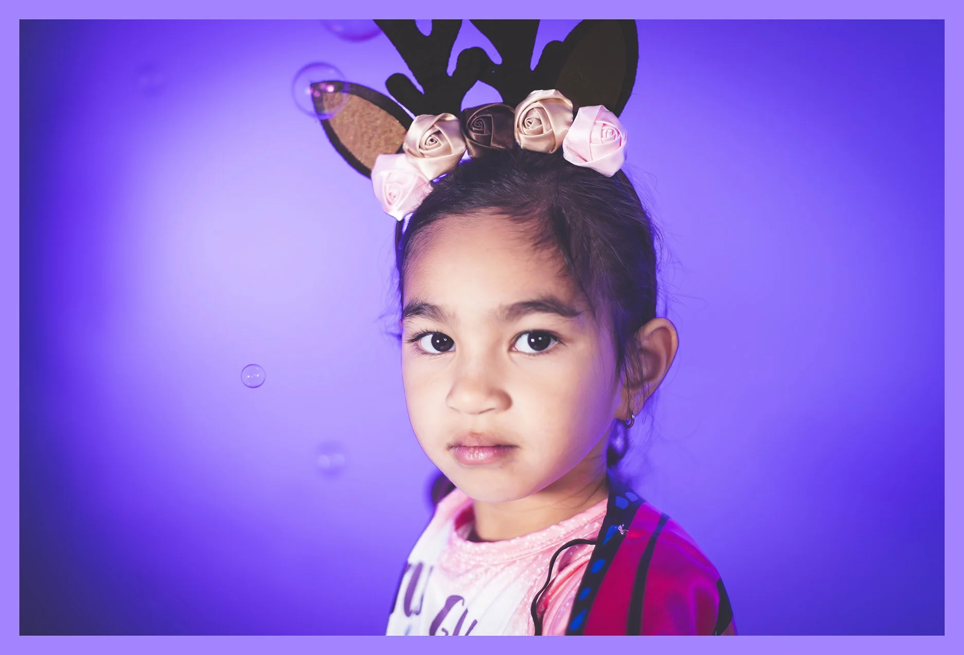 A young girl with dark hair and big eyes wears a headband with attached reindeer antlers and pink and brown flowers. She has a neutral expression and is dressed in a pink and white shirt with a red jacket. The background is purple with small, floatin