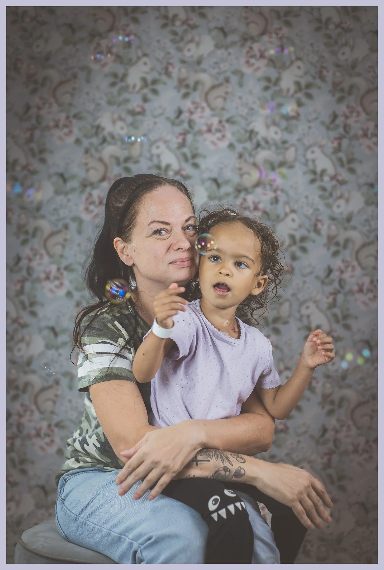 A woman with dark hair and tattoo on her arm sitting on a stool, holding a young girl with curly hair, both looking at bubbles floating around in front of a floral-patterned backdrop.