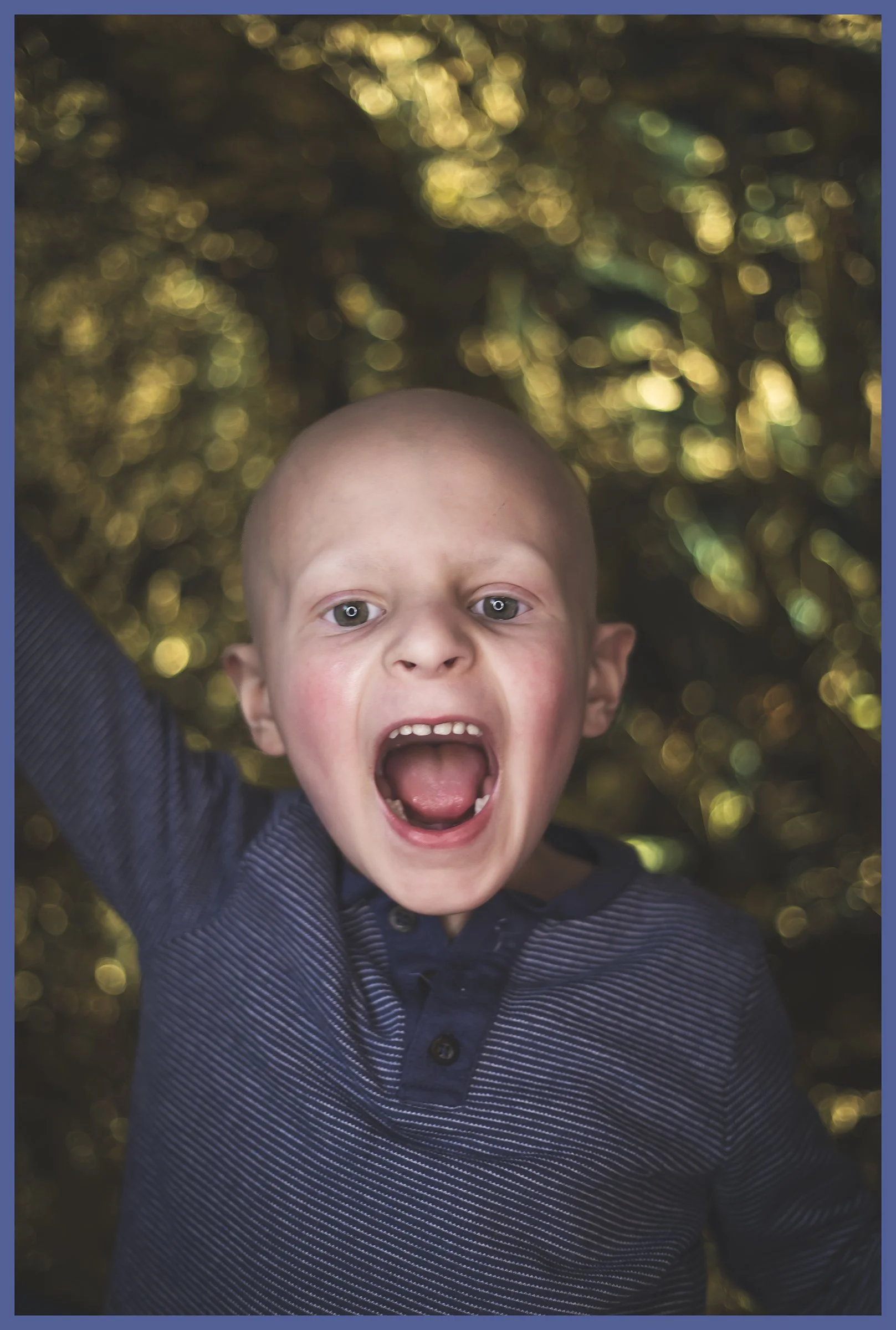 A young boy with a bald head and open mouth, appearing to shout or yell, wearing a dark blue, striped shirt, with a blurred, golden bokeh background.