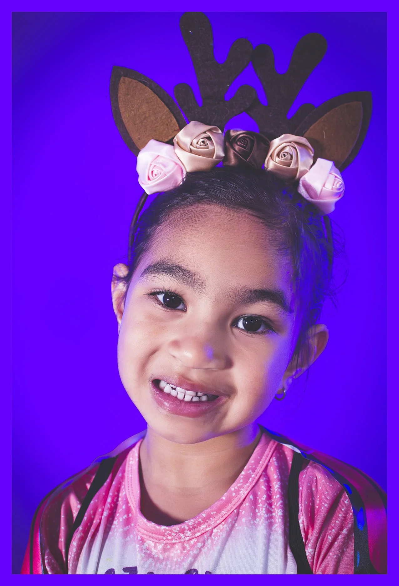 A young girl wearing reindeer antlers headband decorated with pink and gold roses, smiling at the camera with a vibrant blue background.