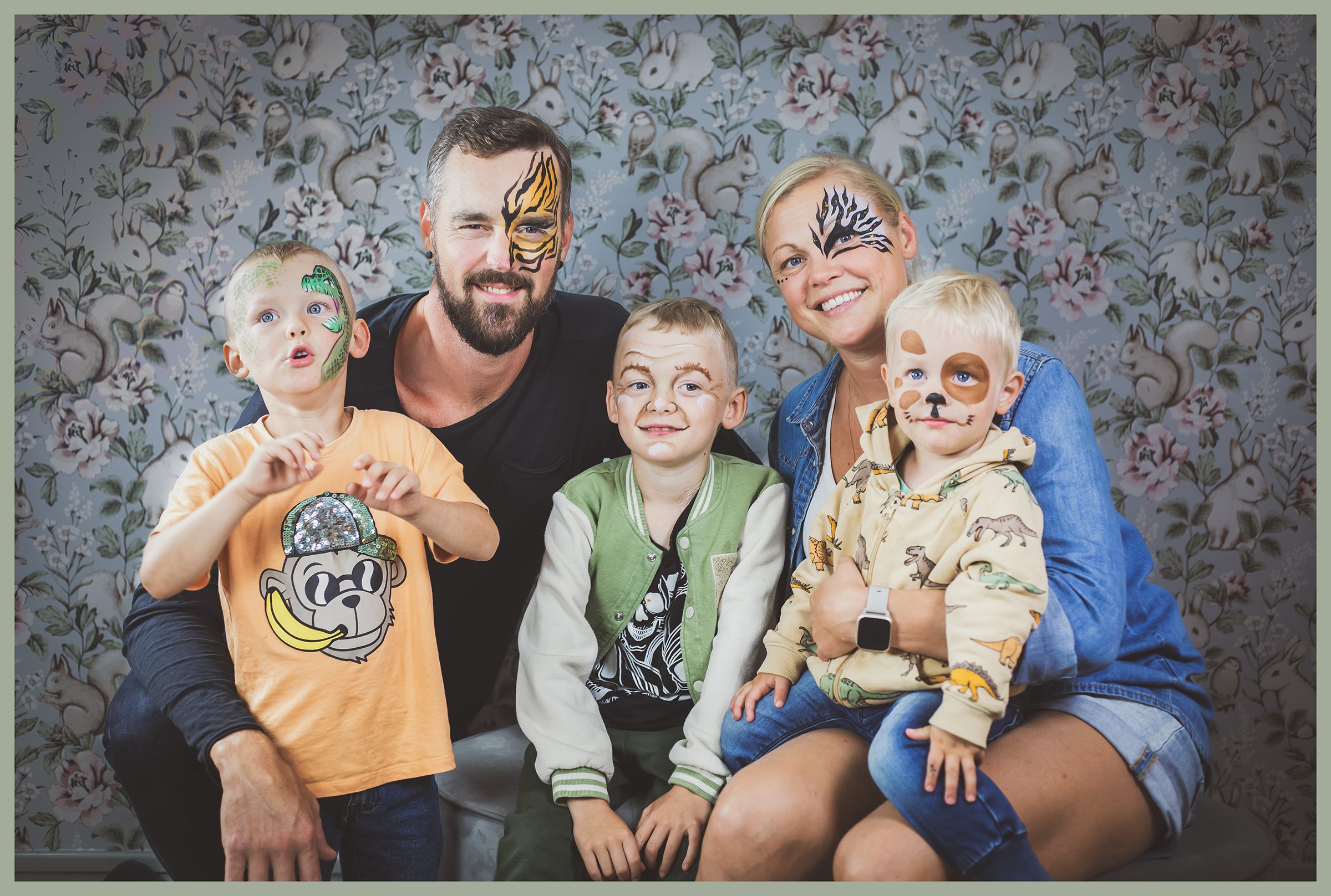 A family of five with face paint and costumes posing together indoors in front of a floral-patterned wallpaper background.
