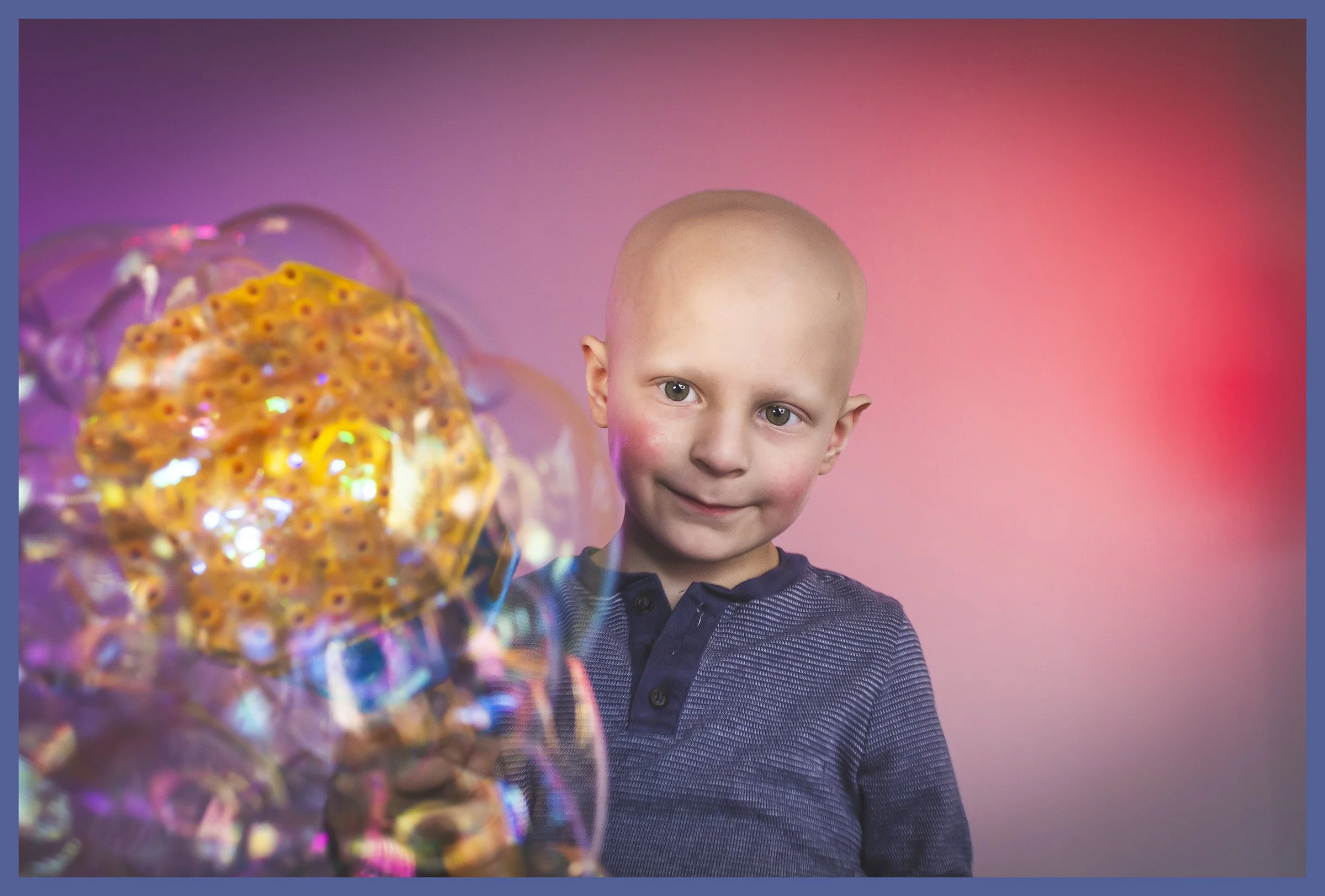 A young bald boy with a slight smile, wearing a dark blue shirt with buttons, is looking at a bubble wand creating iridescent bubbles against a pink and purple background.