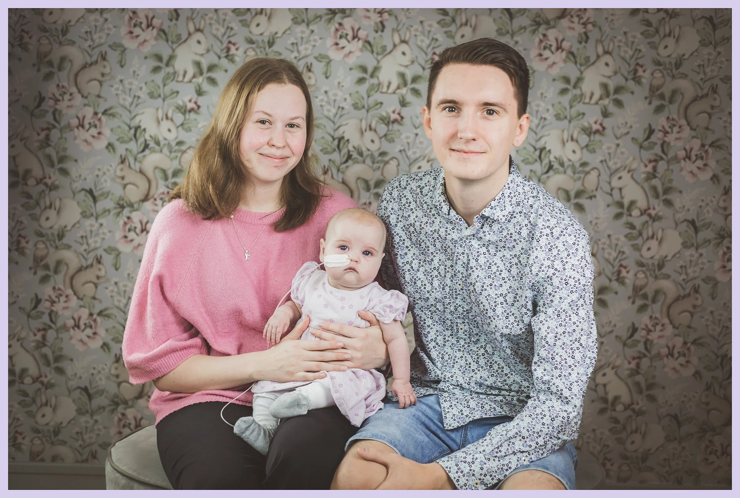 A family of three sitting in front of a floral wallpaper: woman in pink sweater, man in patterned shirt, and a baby girl with medical equipment.