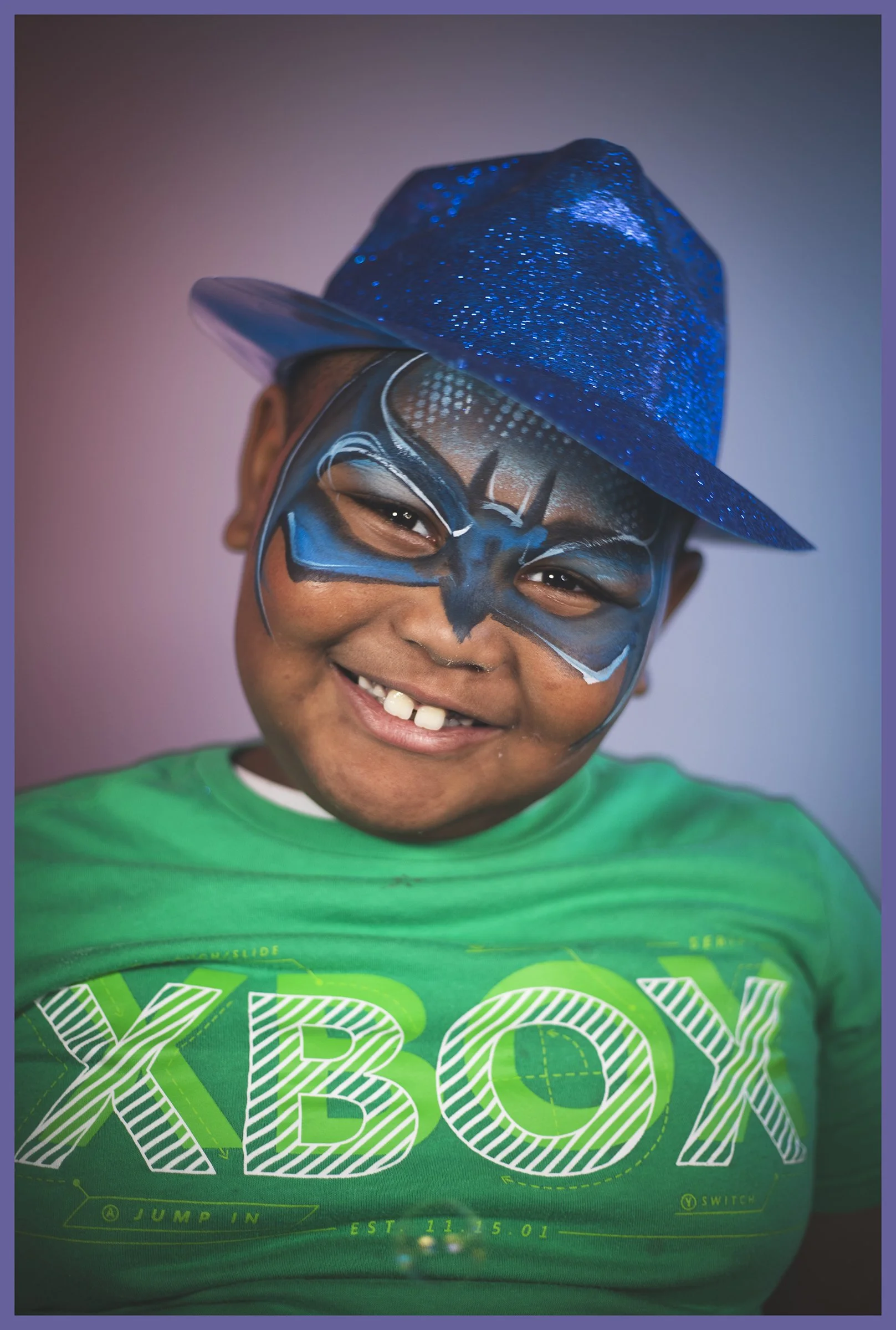 A smiling young boy wearing a blue glittery hat, face paint resembling superhero mask, and a bright green XLBOX T-shirt.