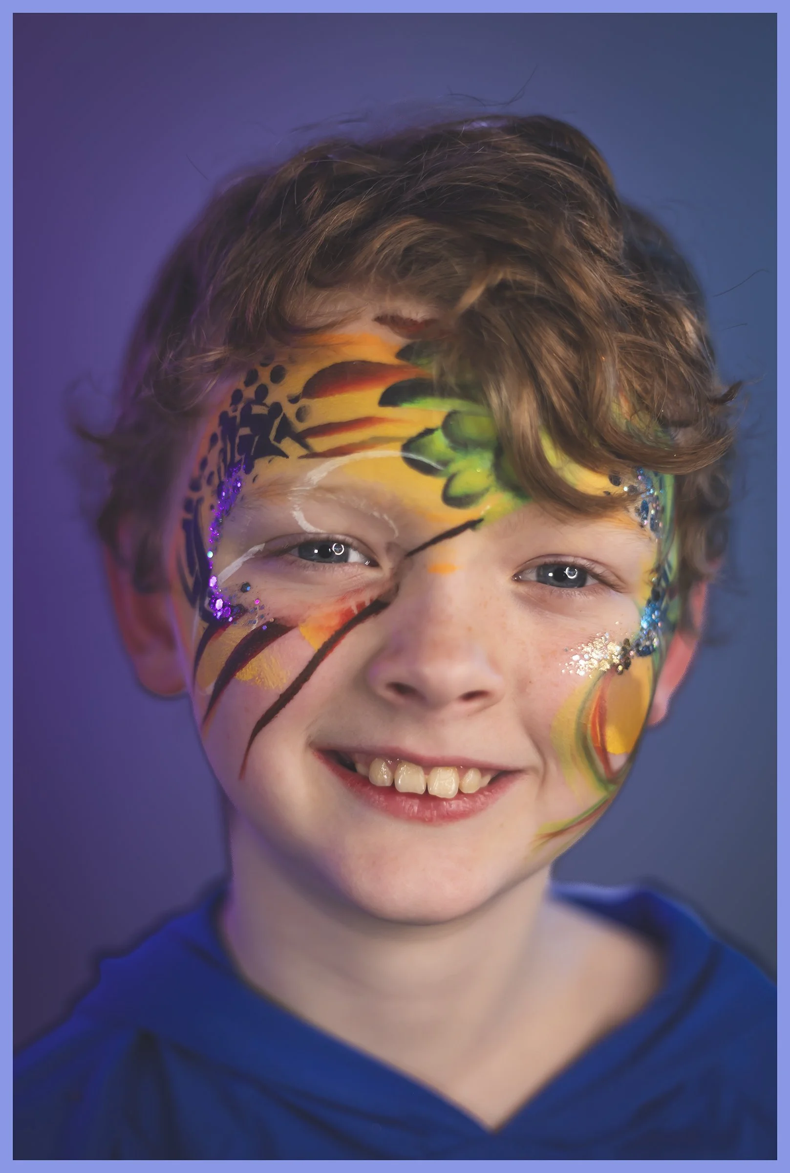 A young boy with colorful face paint, glitter, and a big smile, wearing a blue hoodie.