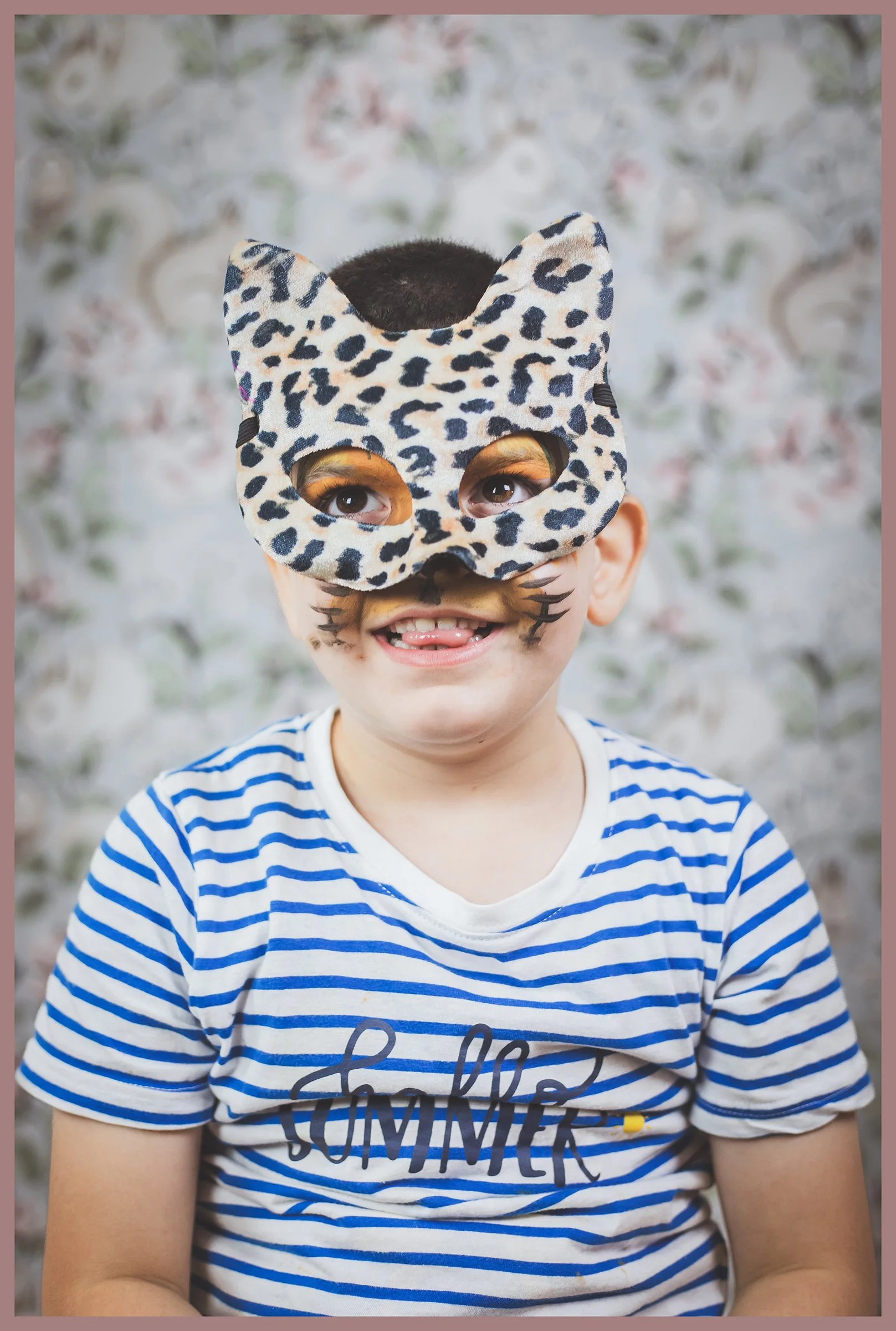 A young boy wearing a leopard-print cat mask, with face paint of whiskers, smiling and sticking out his tongue. He is dressed in a blue and white striped t-shirt with the word 'SUMMER' on it, in front of a floral background.