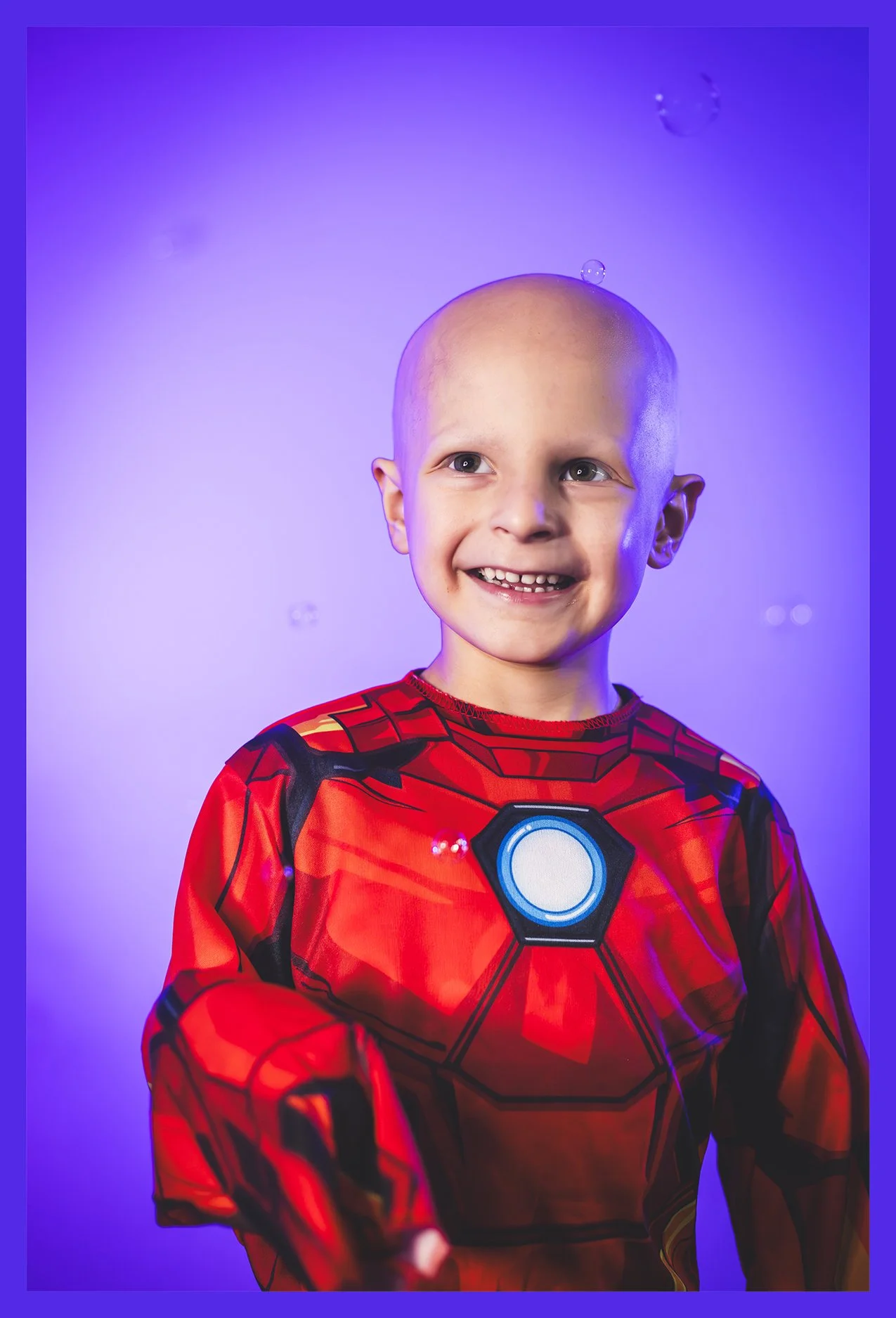 A young boy wearing an Iron Man costume, smiling with missing front teeth, against a purple background with floating bubbles.