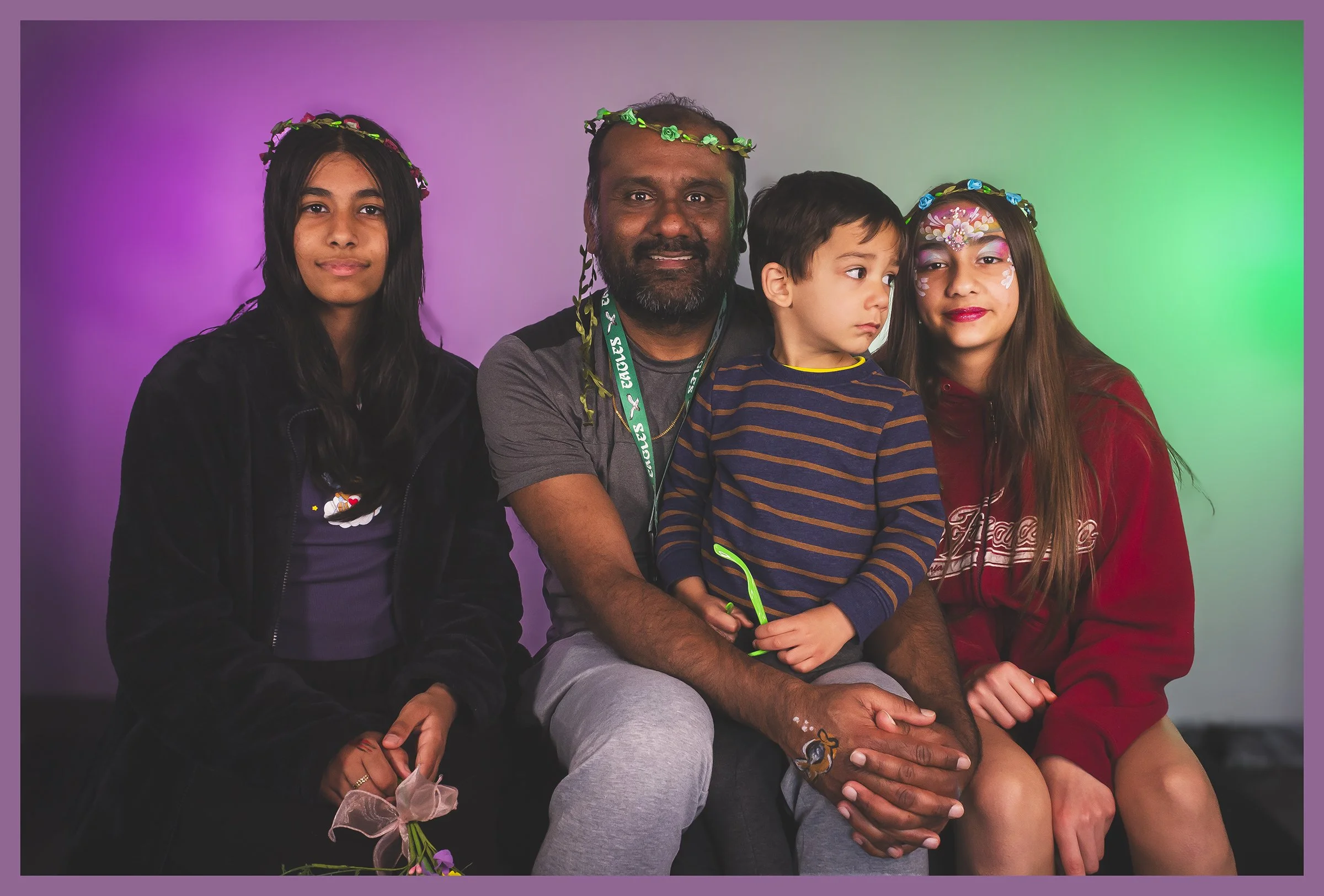 A group of five people, including a man, two young women, and two children, sitting together in front of a purple and green gradient background. Some members are wearing floral headbands and face paint.