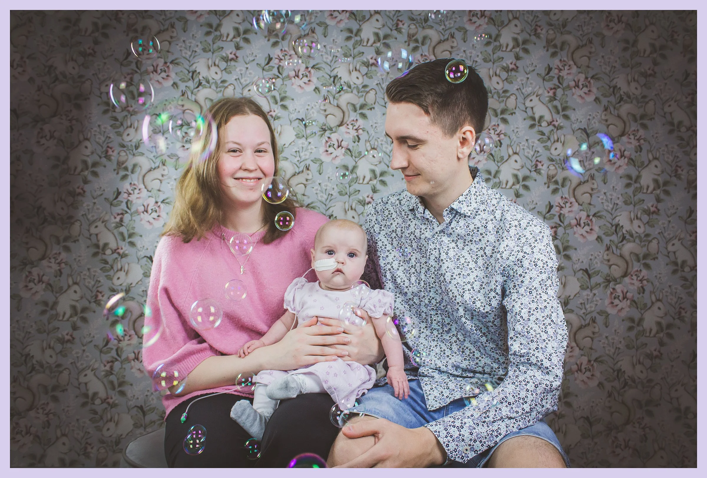 Family portrait with a mother, father, and a baby girl sitting on a bench, surrounded by floating soap bubbles. The mother is wearing a pink sweater and smiling, the father has short dark hair and a patterned shirt, and the baby has a nasal tube, wea