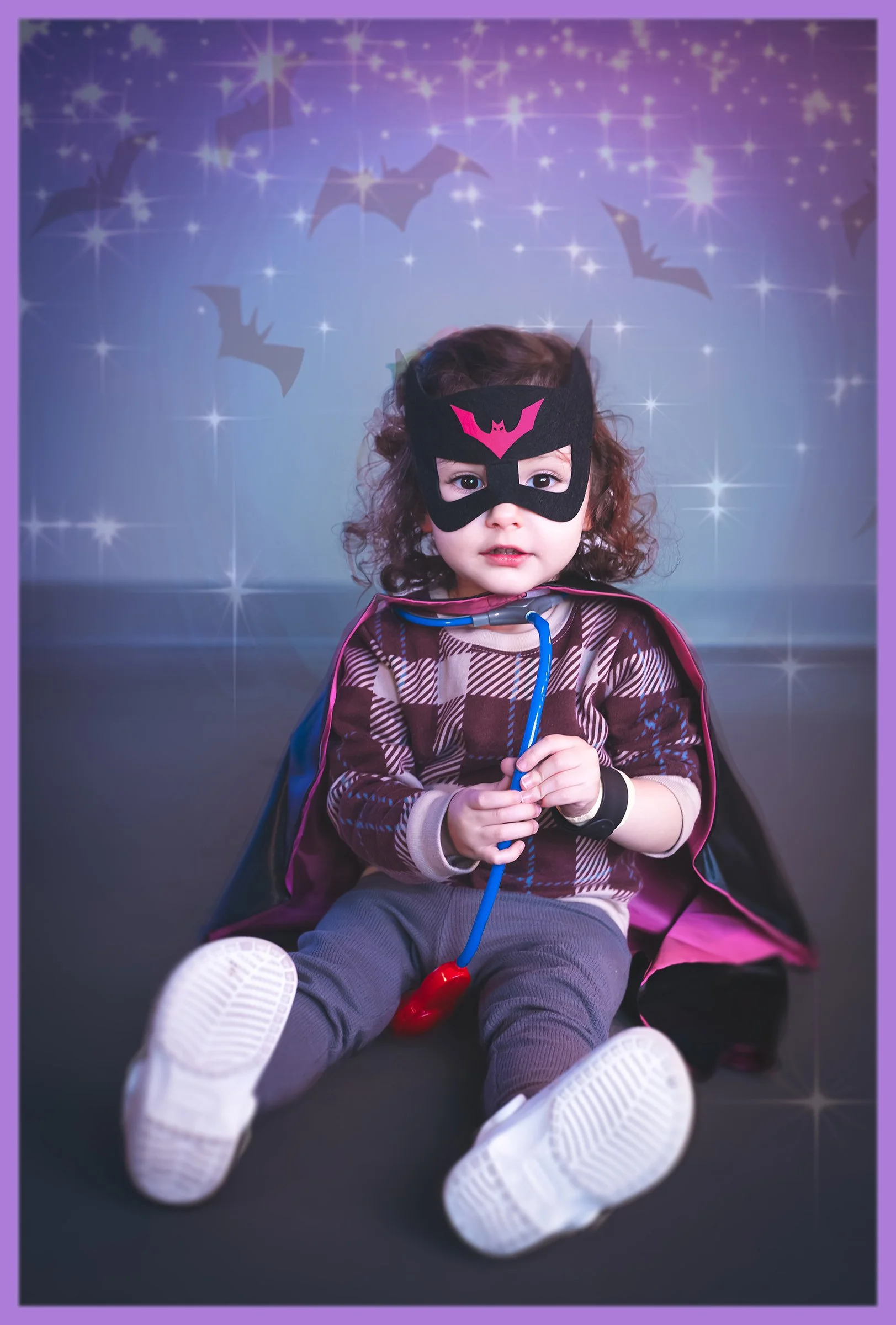 A young child dressed in superhero costume, with a black and pink mask, cape, and holding a toy stethoscope, sitting on the floor in front of a backdrop with bats and stars.