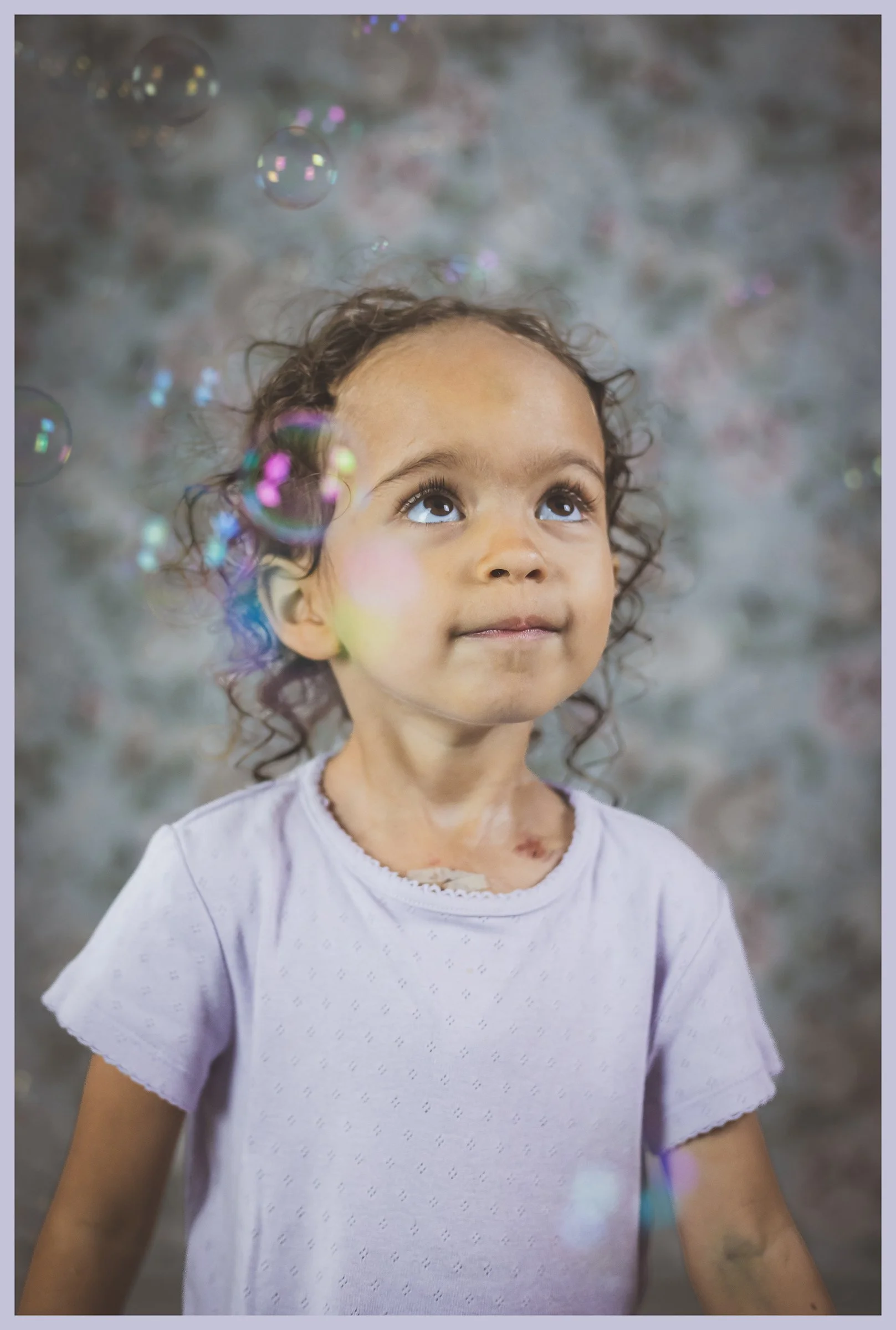 A young girl with curly hair looking up at floating soap bubbles in front of a blurred, colorful background.