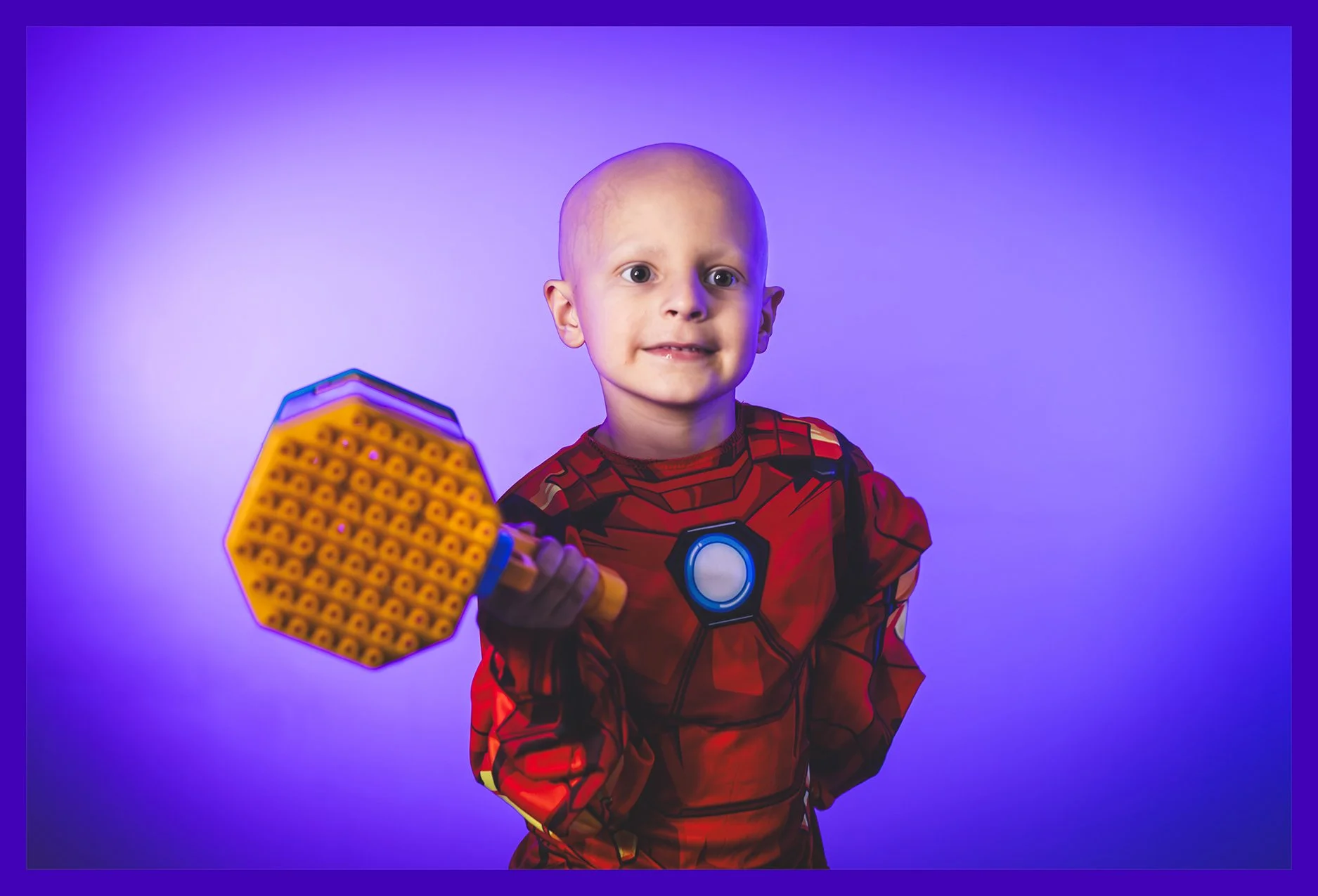 A young boy dressed in a red Iron Man costume, holding a Lego weapon, against a purple gradient background.