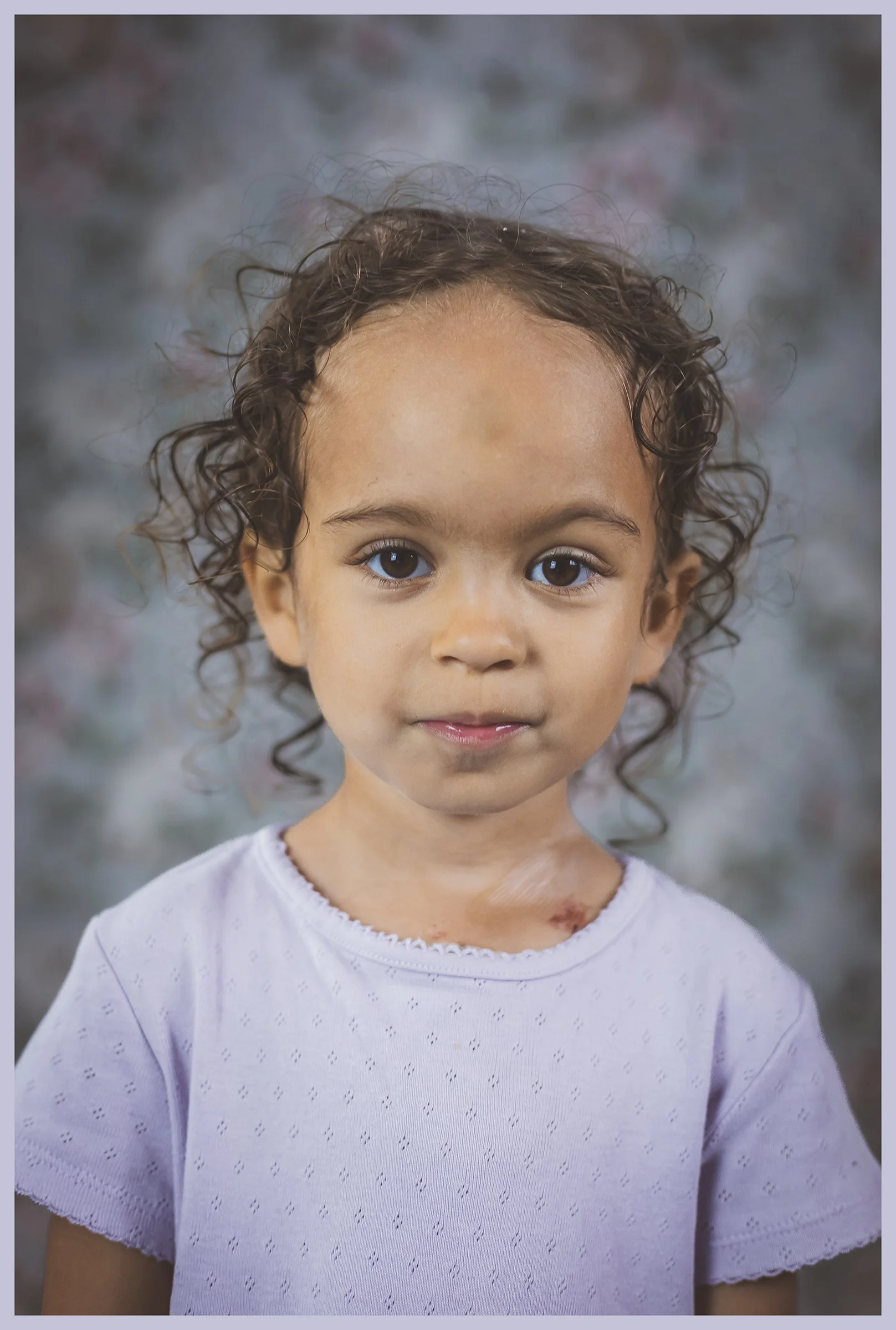 A young girl with curly hair, large brown eyes, and tan skin, wearing a white shirt, standing in front of a blurred floral background.