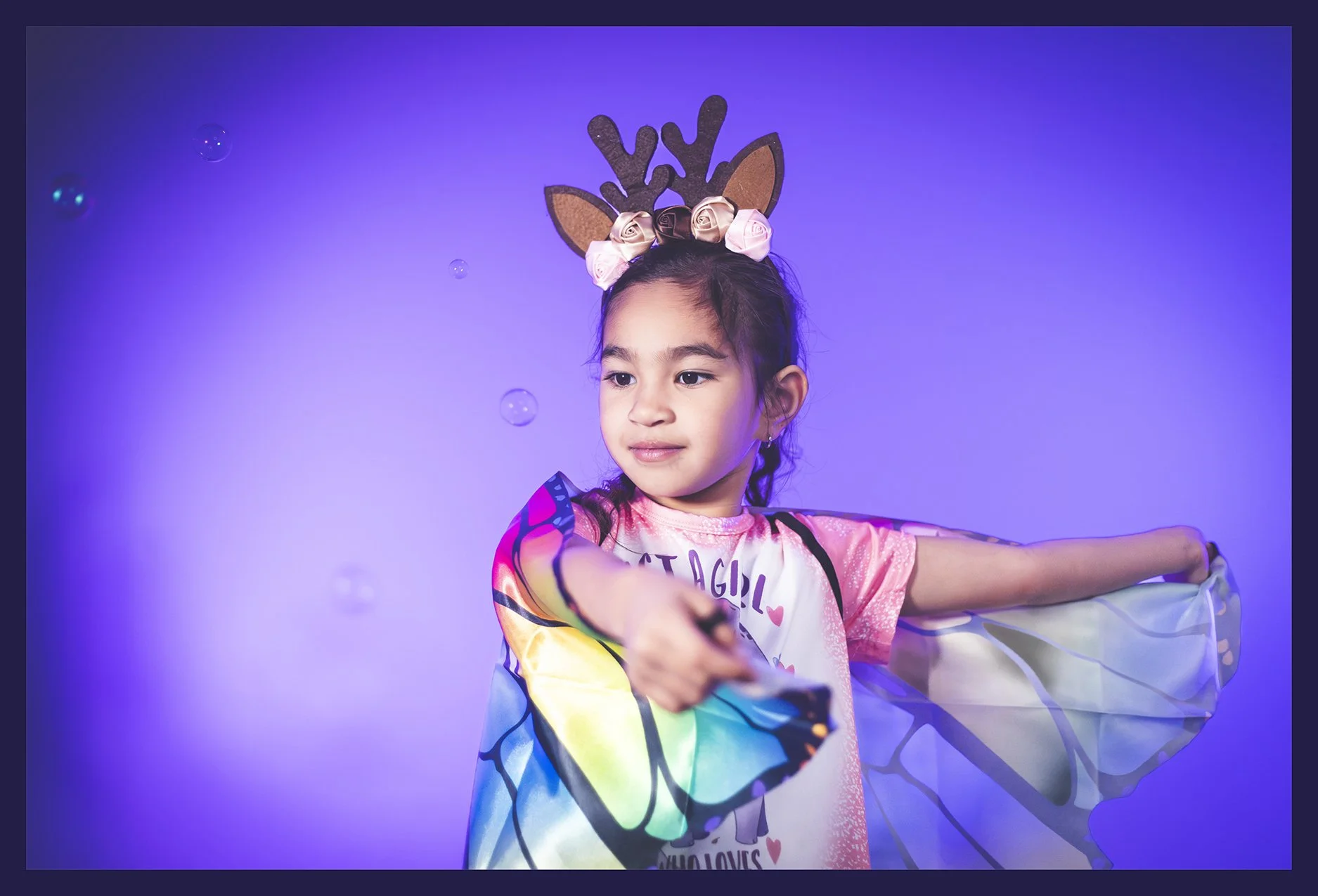 A young girl wearing reindeer antler headband with pink roses, extending her right arm holding a remote, against a purple background with floating bubbles.