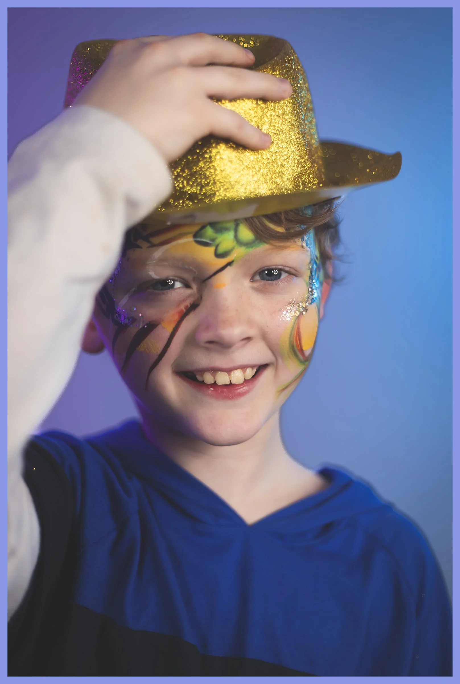 A smiling boy with face paint and glitter, wearing a blue shirt, holds a shiny gold hat.