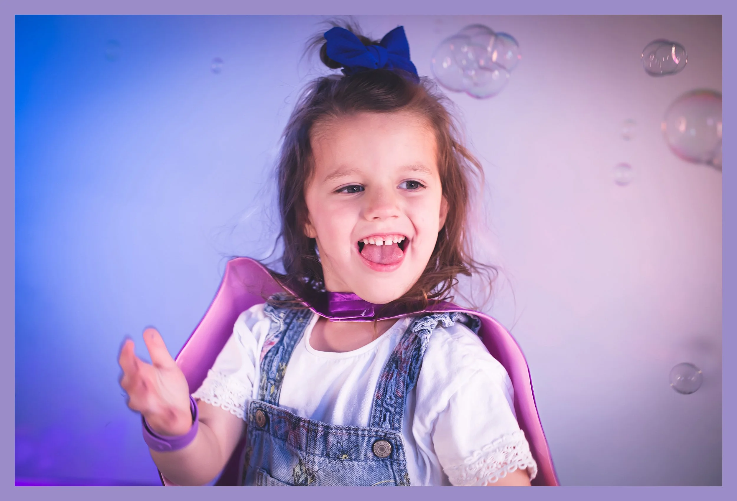 Young girl with curly brown hair tied with a blue bow, wearing a white lace-trimmed shirt and denim overalls, smiling and waving, surrounded by floating soap bubbles against a pastel-colored background.