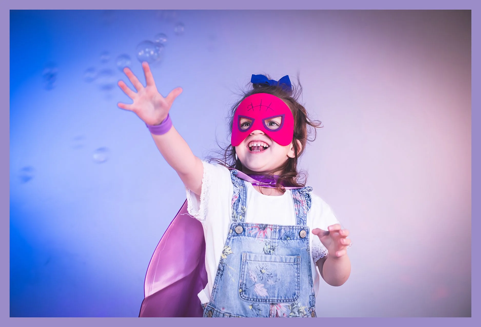 A smiling young girl wearing a pink superhero mask, a blue bow headband, a white shirt, denim overalls, and a pink cape, reaching out towards bubbles against a gradient background.