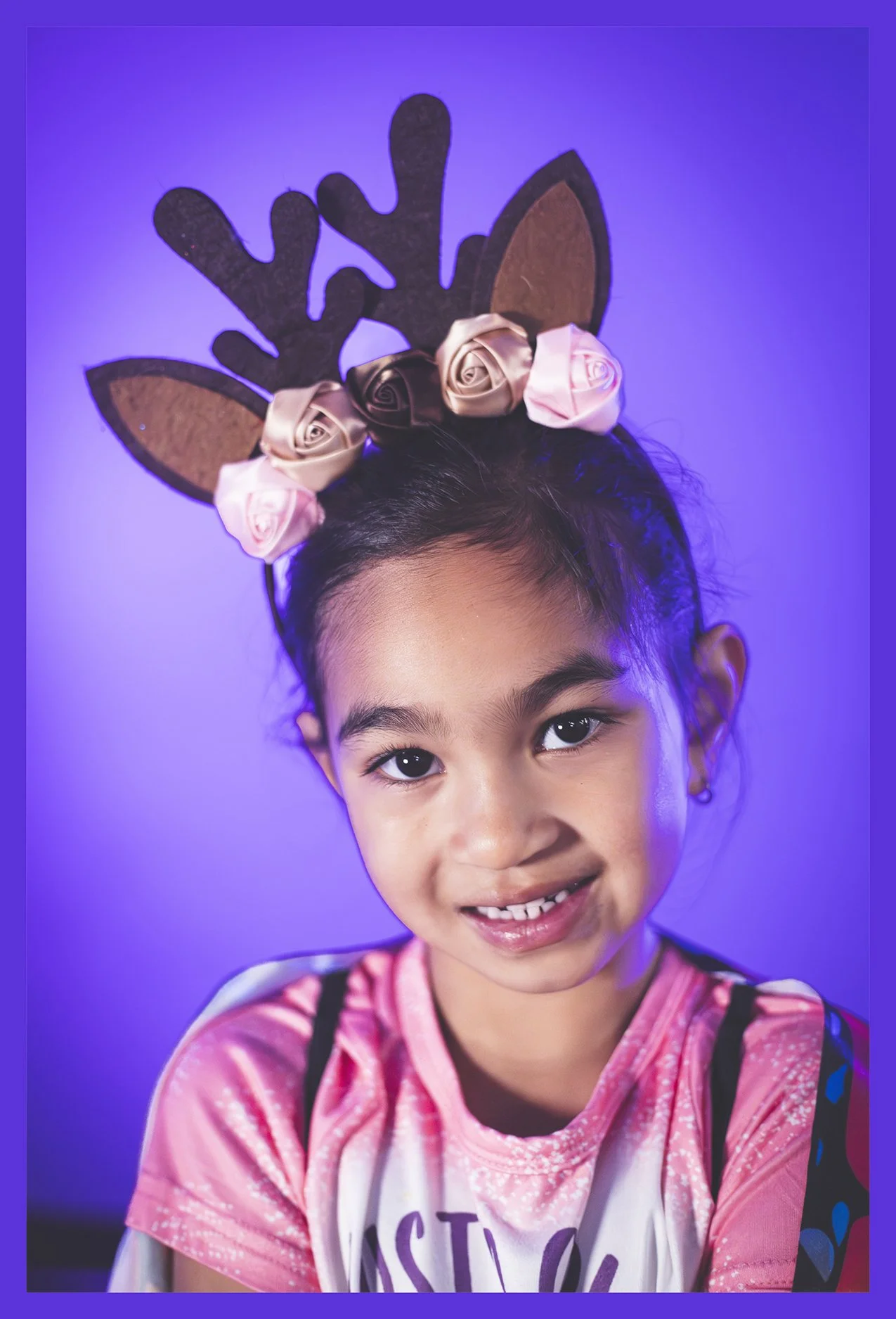 A young girl wearing a pink shirt with black shoulder straps, smiling, with reindeer antlers headband decorated with pink and beige roses against a purple background.