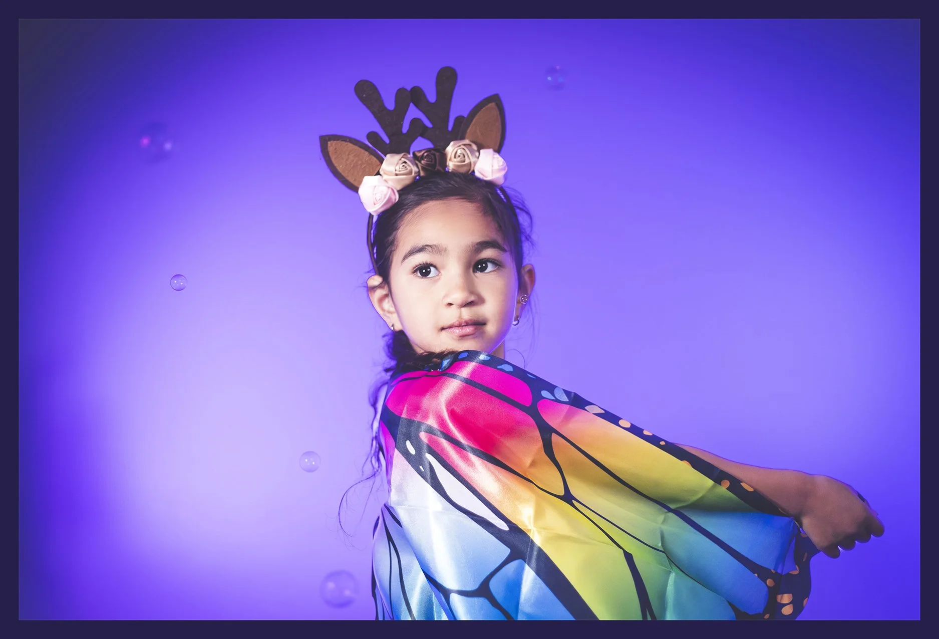 Young girl wearing reindeer antler headband with pink and white roses, wrapped in a colorful butterfly or fairy wing costume, against a purple background with floating bubbles.