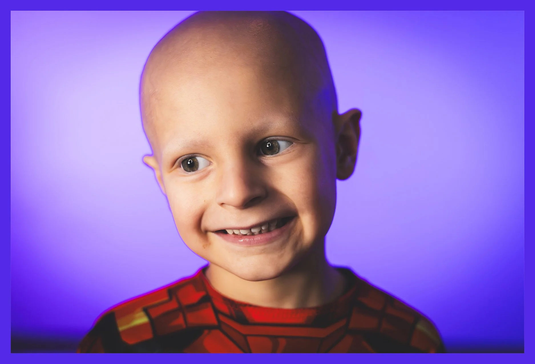 A young boy with a bald head smiling and looking to the side, wearing a red and black Spider-Man shirt, against a purple background.