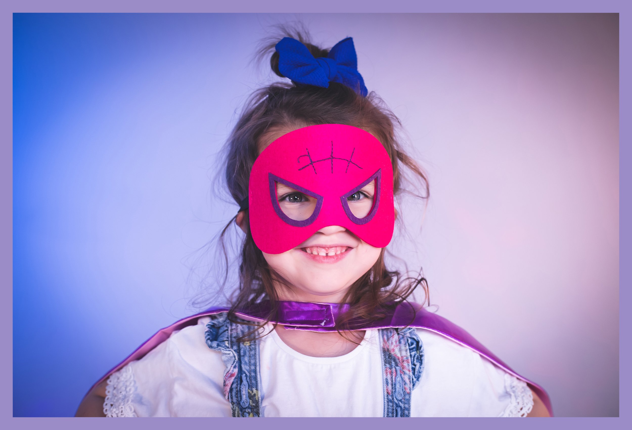 Young girl wearing a pink and purple Spider-Man mask, a blue bow in her hair, a purple cape, and a white dress with lace details, smiling at the camera.