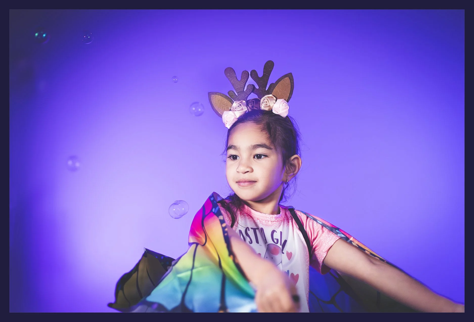 A young girl wearing a reindeer headband with pink roses and rainbow-colored wings, smiling softly and reaching out her arm, against a purple background with small bubbles.