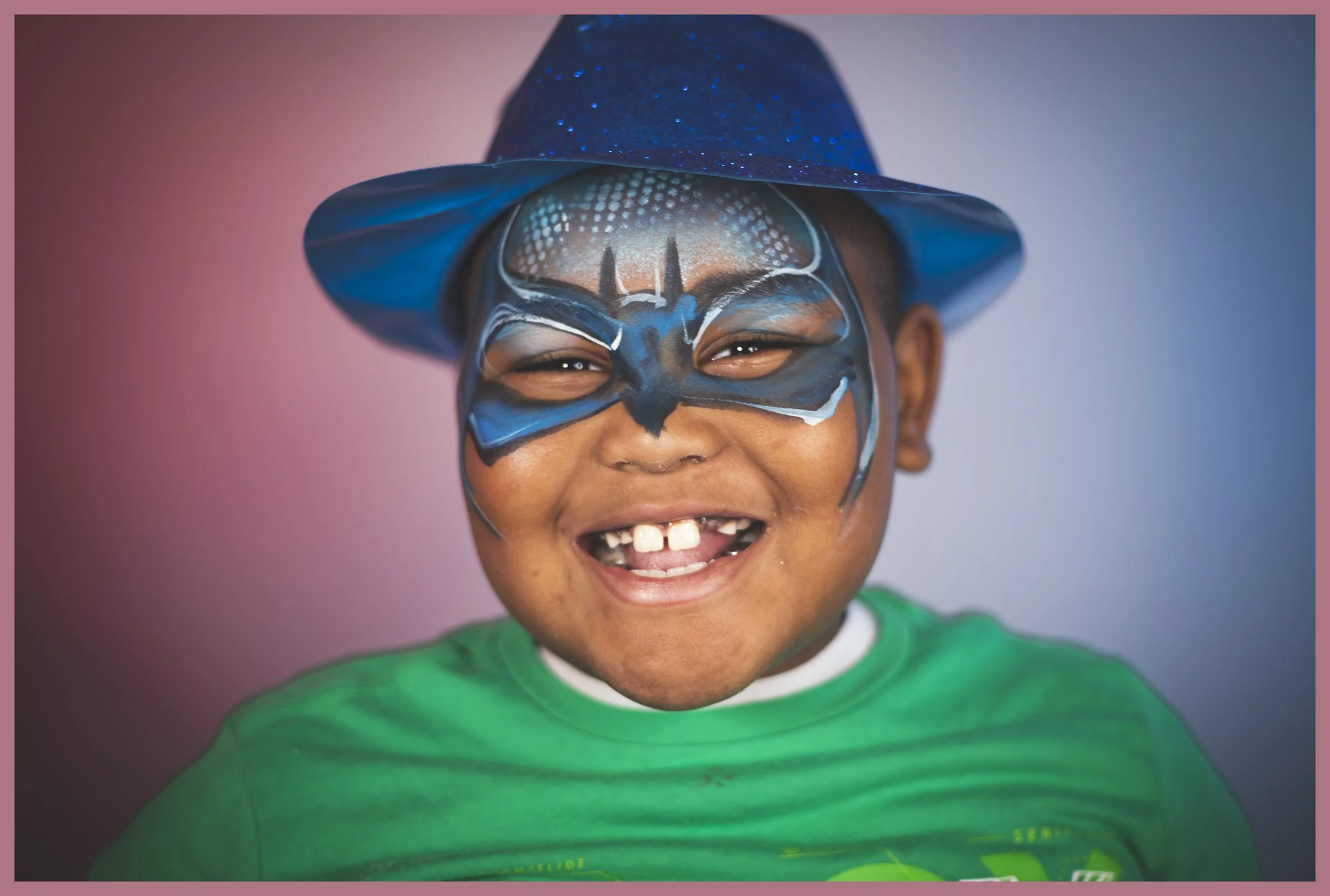 A smiling young boy with painted face as a blue butterfly and wearing a blue hat. He is dressed in a green shirt.