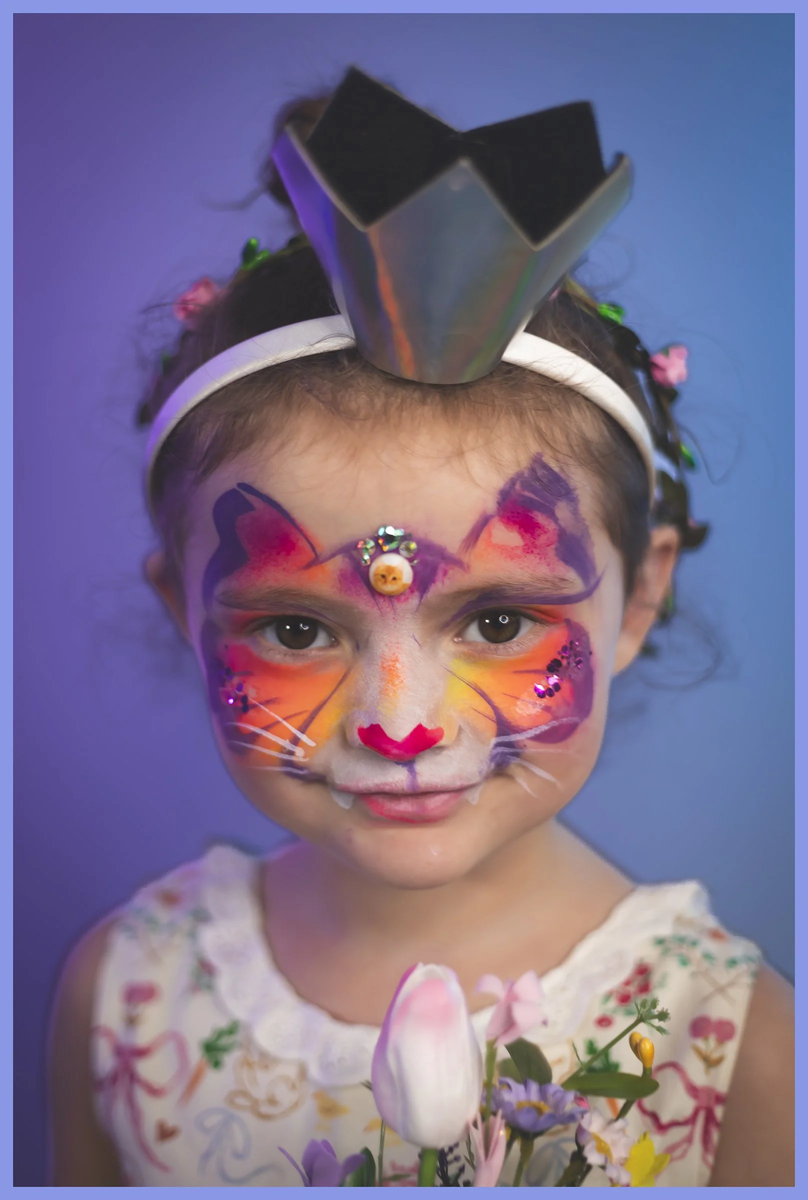 A young girl with face paint resembling a colorful butterfly, wearing a silver crown headband, surrounded by brightly colored flowers. She is smiling softly against a blue background.