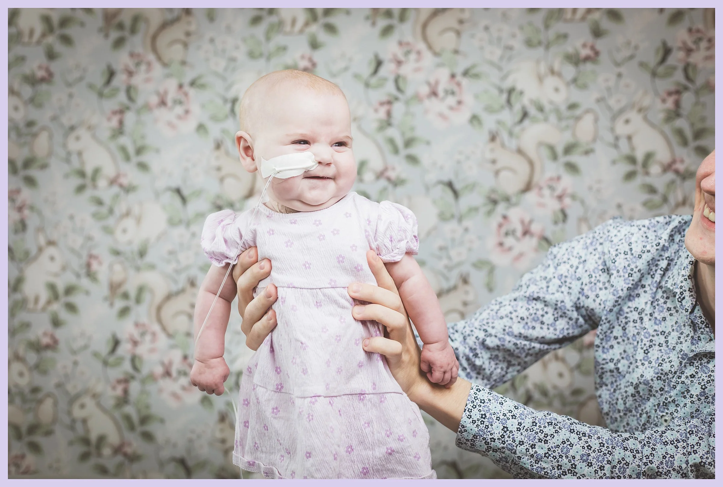 A smiling baby girl with a hospital nasal oxygen tube, being held up by an adult, with a colorful floral wallpaper background.