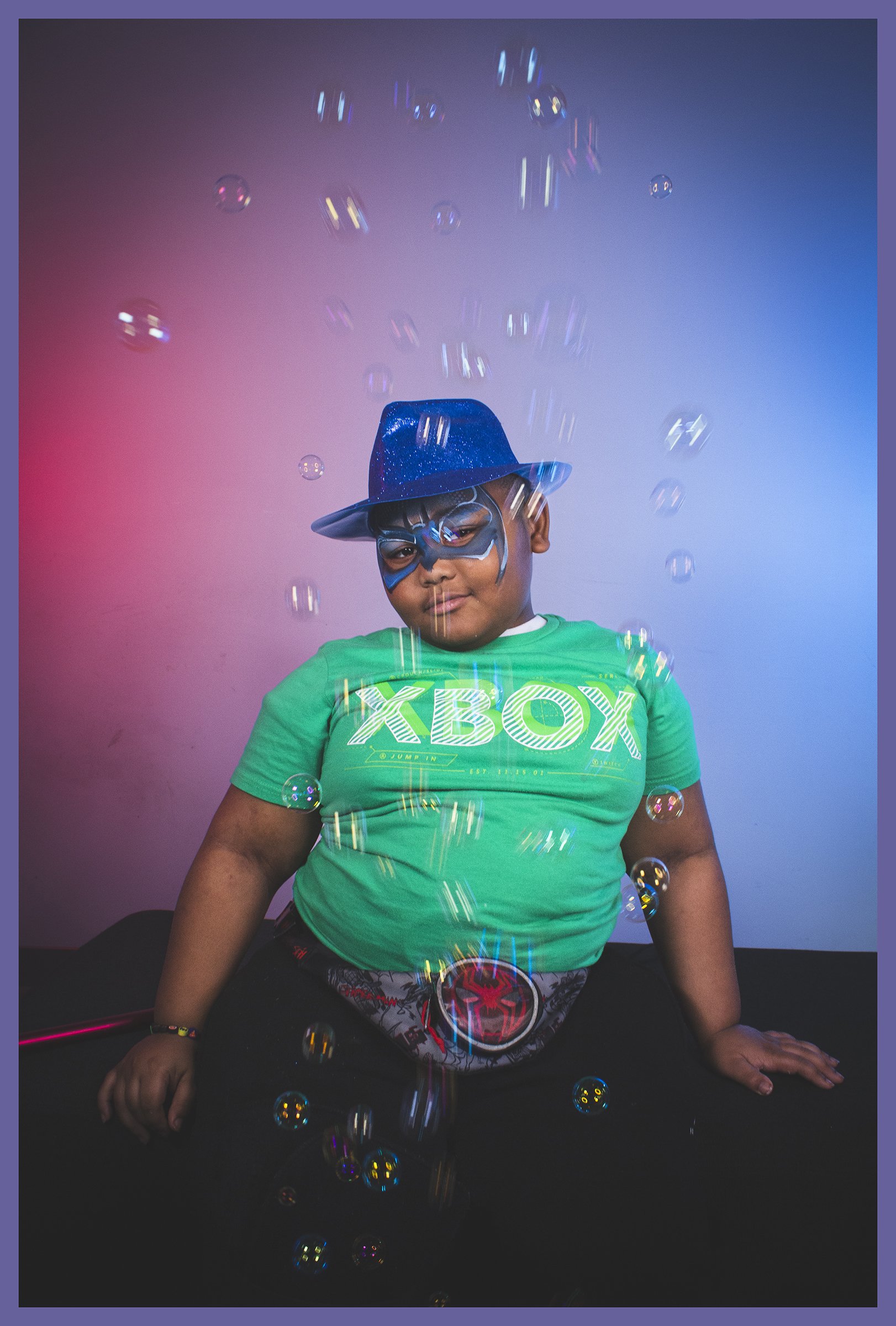 A young boy dressed in a green 'XBOX' shirt, wearing a blue wizard hat and a blue superhero mask, sits on a black surface surrounded by colorful bubbles and a magical effect with pink and blue lighting.