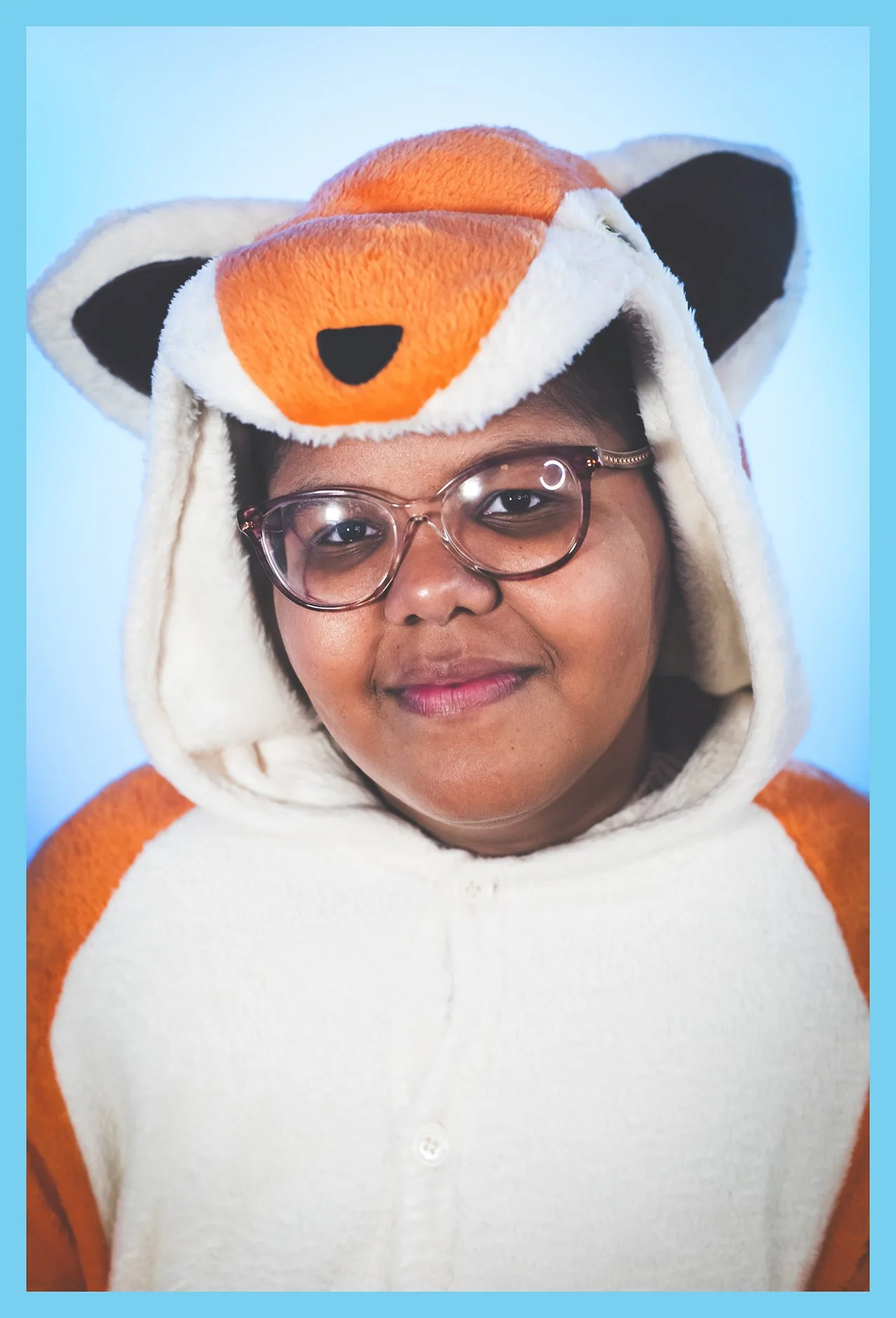 Person wearing a plush fox costume hoodie with the hood over their head, glasses, and smiling at the camera against a light blue background.