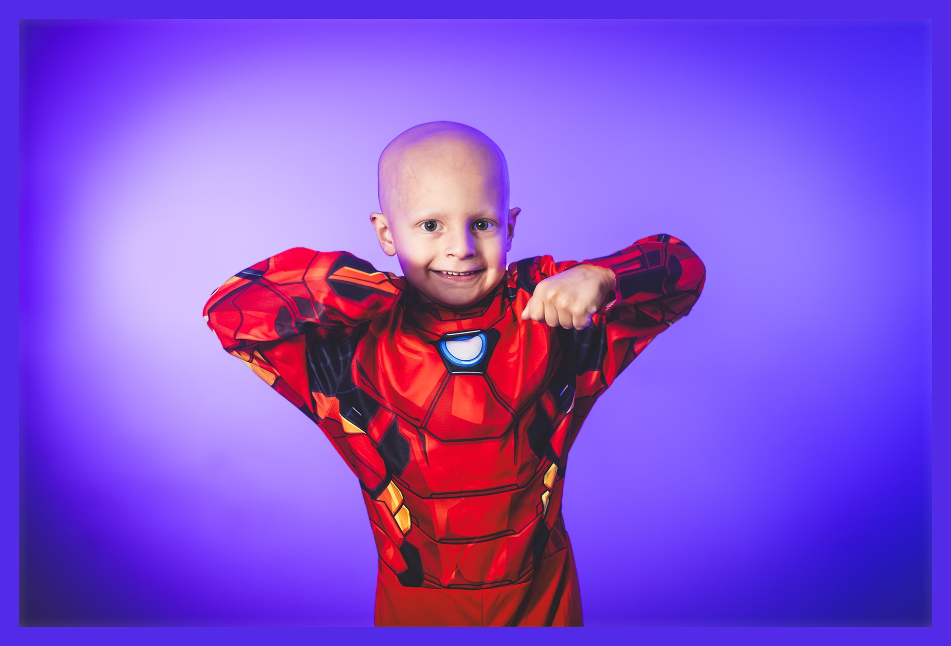 Child dressed in Iron Man superhero costume smiling and showing muscles with purple background.
