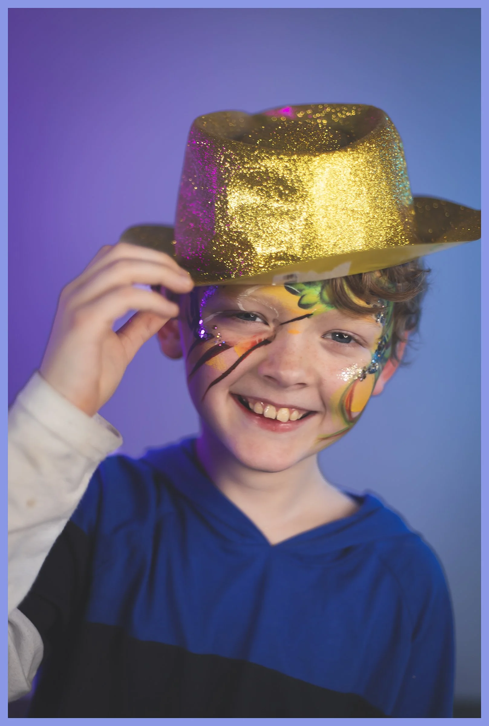 A smiling boy with face paint and glitter, wearing a blue shirt and a gold glittery hat, holding the hat with his right hand.