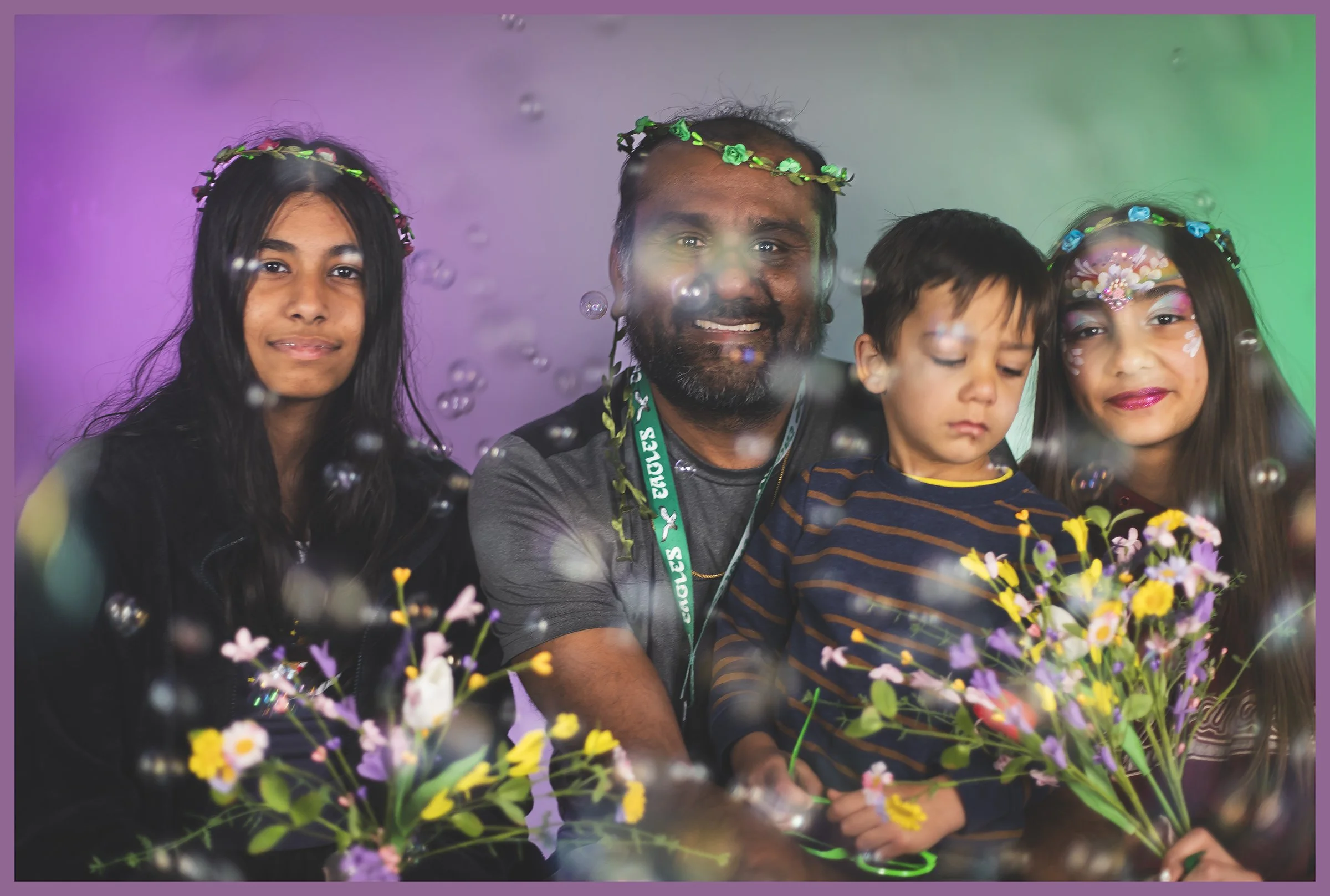 A family of four, two women, a man, and a young boy, sitting together in front of a colorful background with bubbles and holding bouquets of flowers. The women have floral face paint and headbands, smiling, while the boy has a neutral expression.