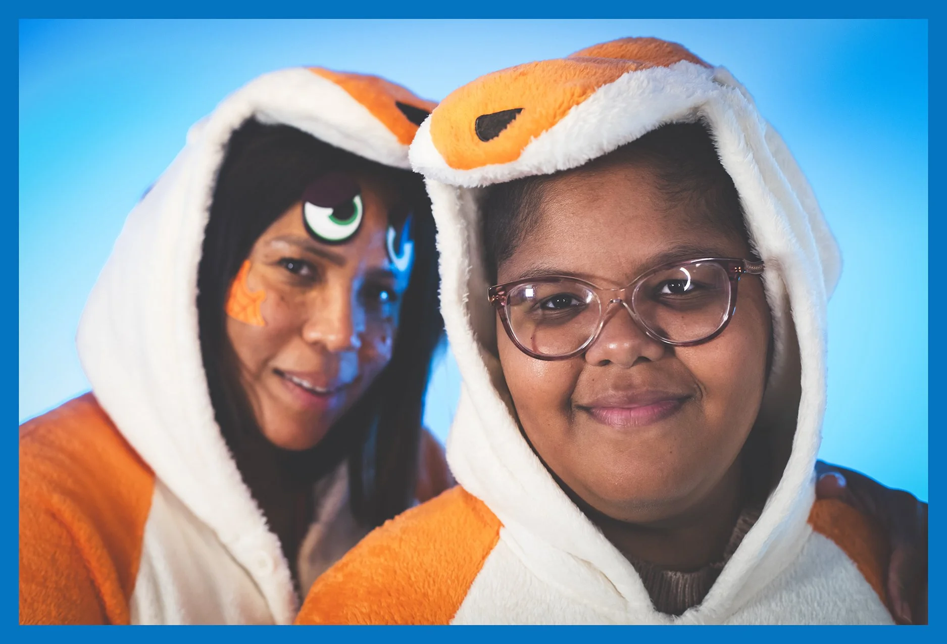 Two people dressed in plush animal costumes resembling a fox and a tiger, wearing hooded onesies with animal faces on the hoods, smiling at the camera against a light blue background.
